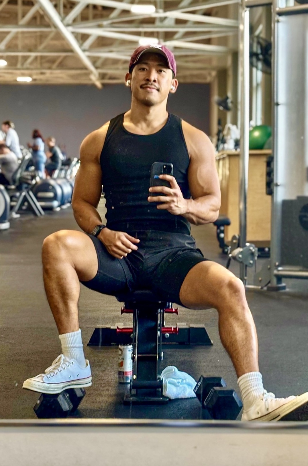 A man takes a selfie at the gym, showcasing his muscular physique. He's wearing a tank top and shorts, with sneakers and a cap.