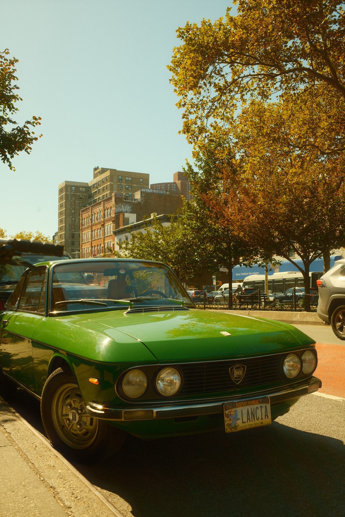 A vintage green Lancia car parked on a city street. The photo captures the car's classic design against a backdrop of buildings and trees.