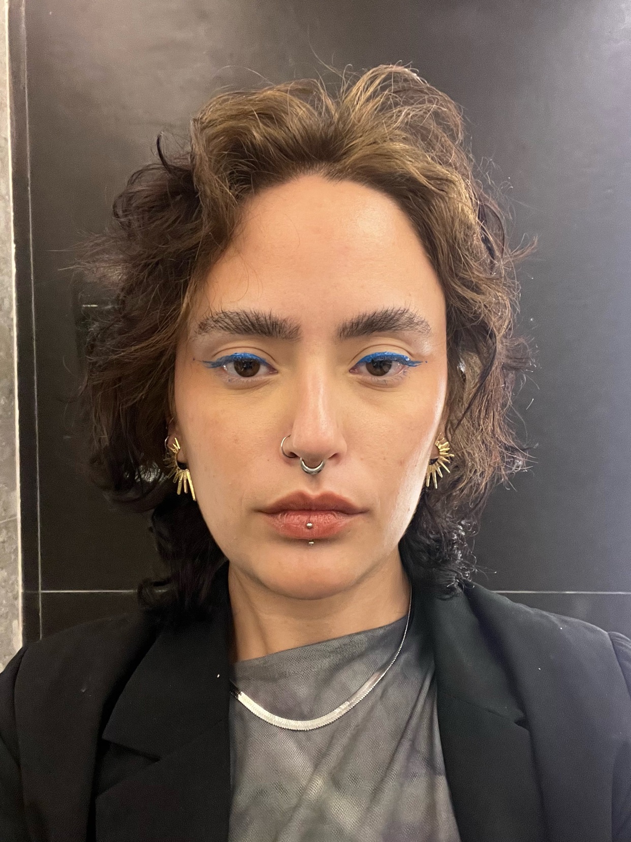 A close-up selfie of a woman with dark, curly hair and striking blue eyeliner. She wears a silver necklace and a black blazer, looking directly at the camera.