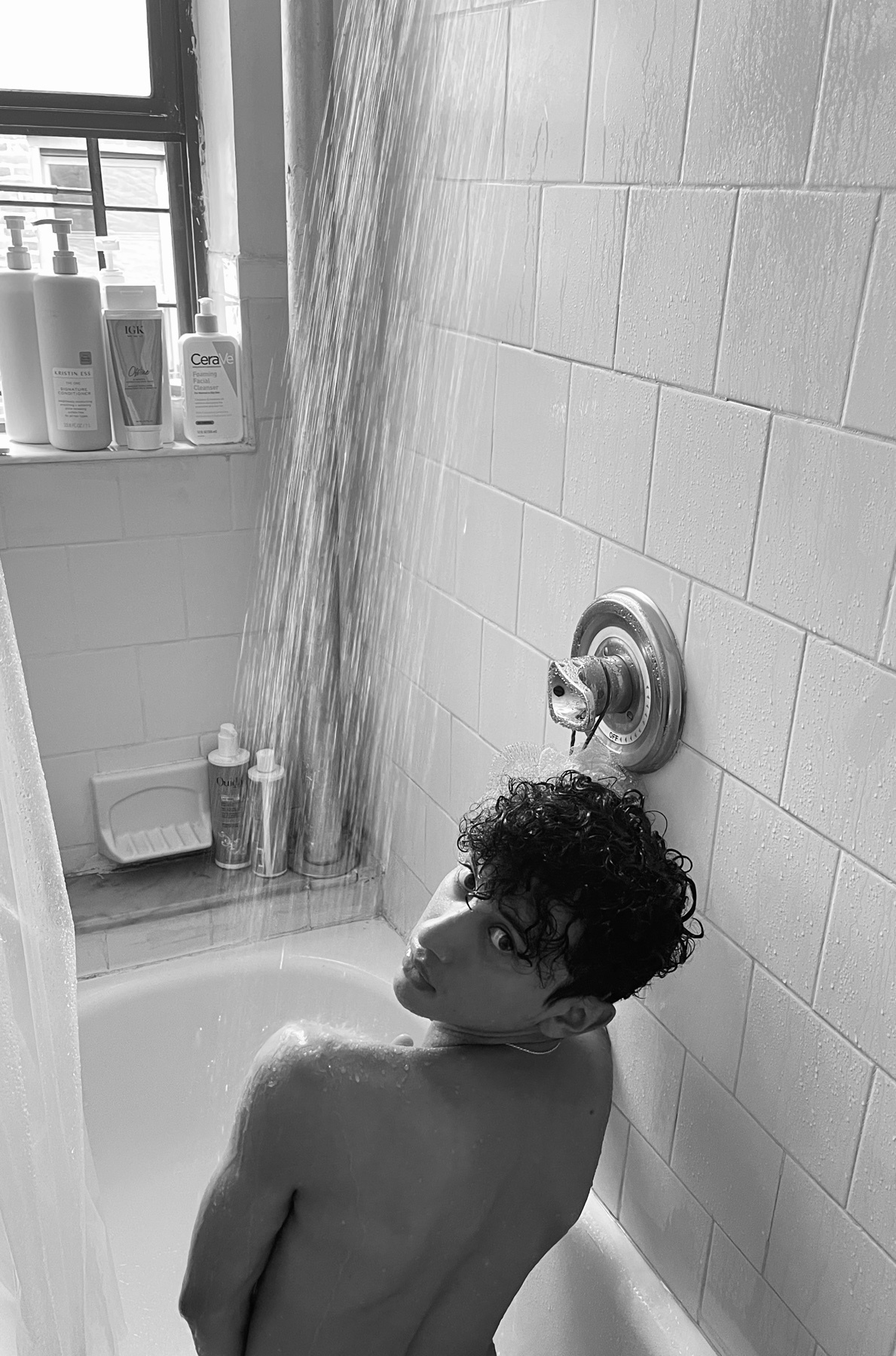 A black and white photo of a man in a shower. He is shirtless and looking over his shoulder.