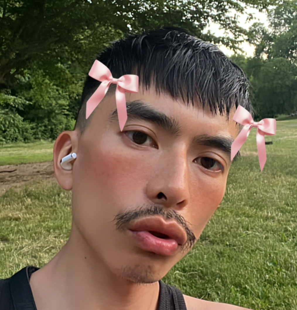 A selfie of a man with a mustache and bows on his face. He is wearing earbuds and has blush on his cheeks.