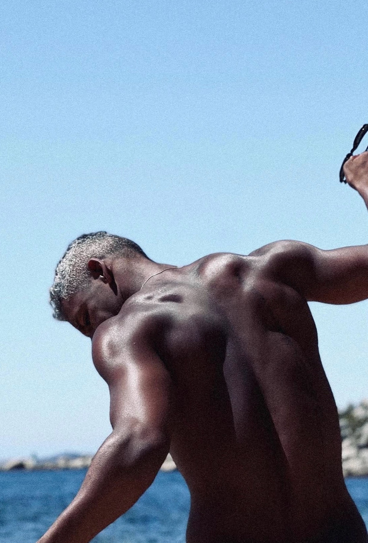 A man with silver hair is captured from behind, showcasing his muscular back and shoulders against a bright blue sky and ocean backdrop. He appears to be holding sunglasses in his right hand.