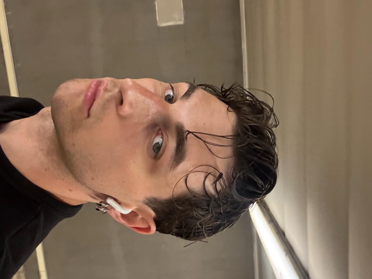 A man poses for a selfie, showcasing his dark, curly hair and modern style. The portrait highlights his features and accessories.