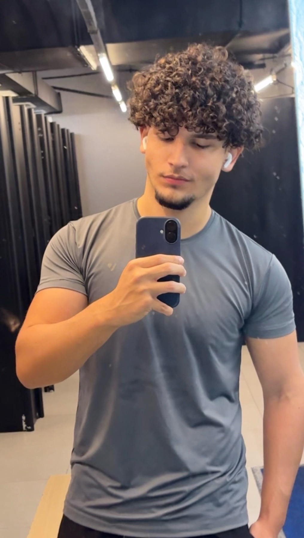 A young man with curly hair takes a selfie in a gym locker room. He is wearing a grey t-shirt and earbuds.