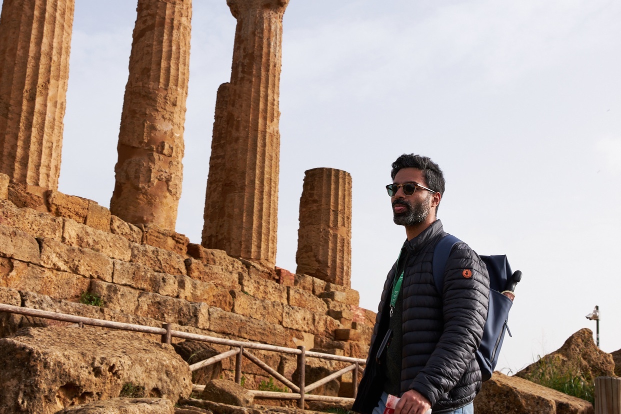 A man with a beard and sunglasses stands in front of ancient Greek ruins with large columns. He is wearing a puffer jacket and a backpack, suggesting he is a tourist exploring a historical site.