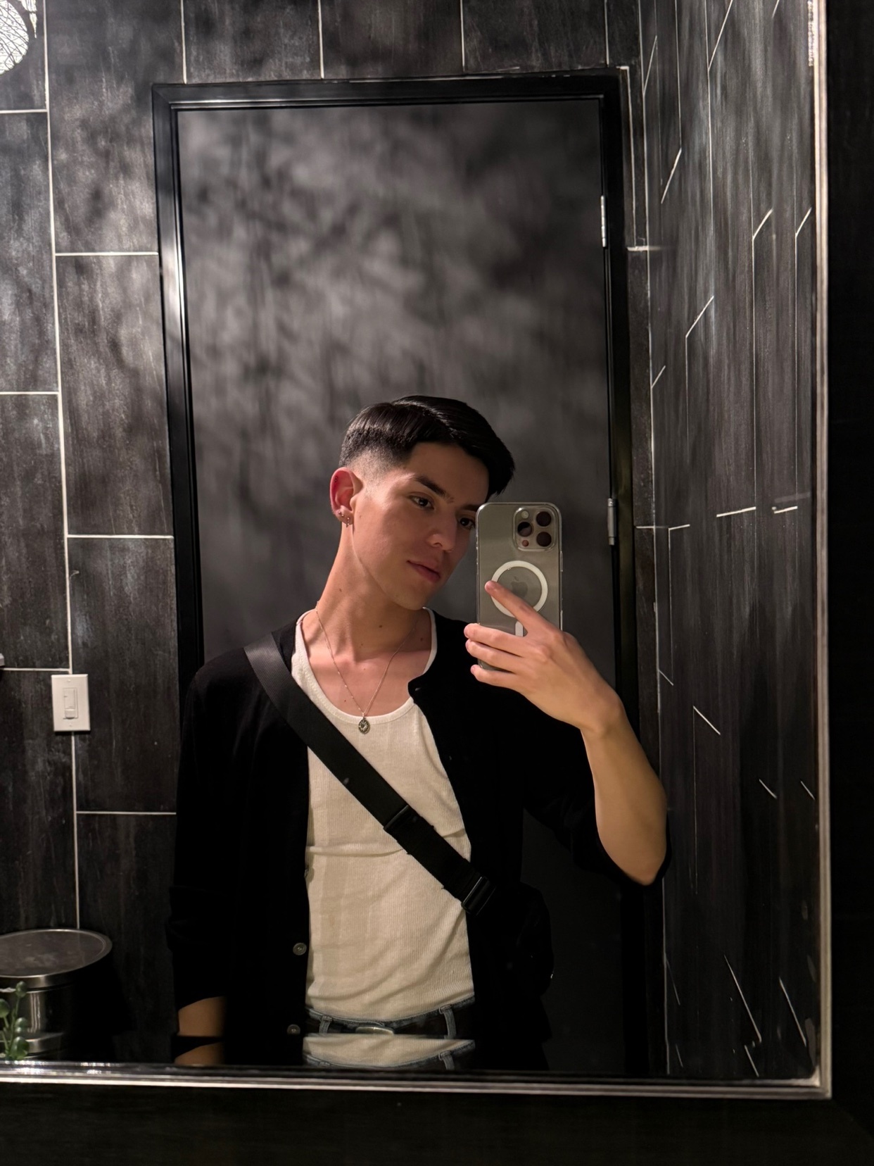 A man takes a mirror selfie in a bathroom. He is wearing a white t-shirt and a black cardigan.