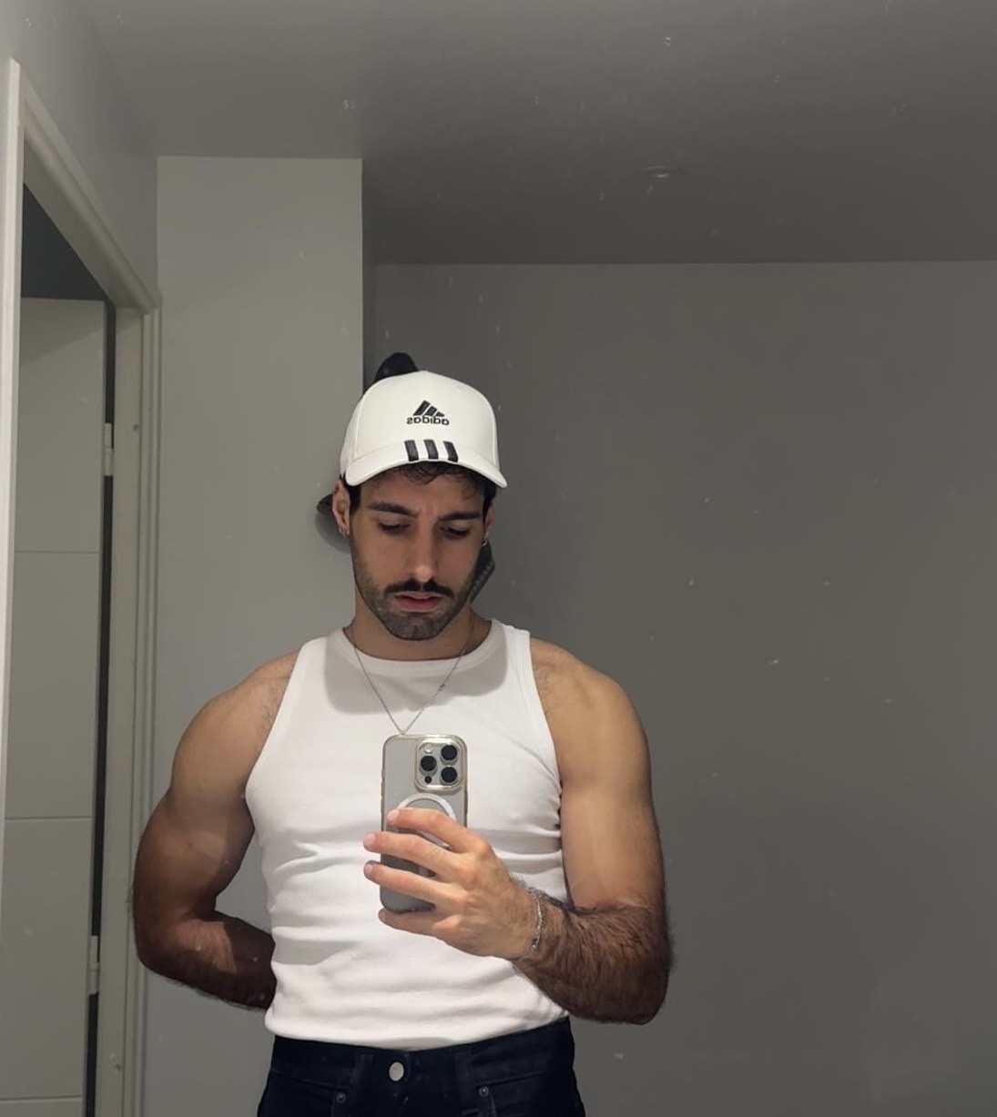 A man in a white tank top and a white baseball cap takes a mirror selfie. He is flexing his arm and has stubble and a moustache.