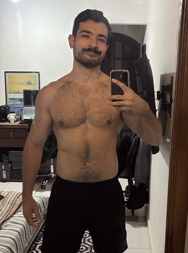 A man with stubble and a moustache takes a mirror selfie indoors. He is shirtless and wearing black shorts.