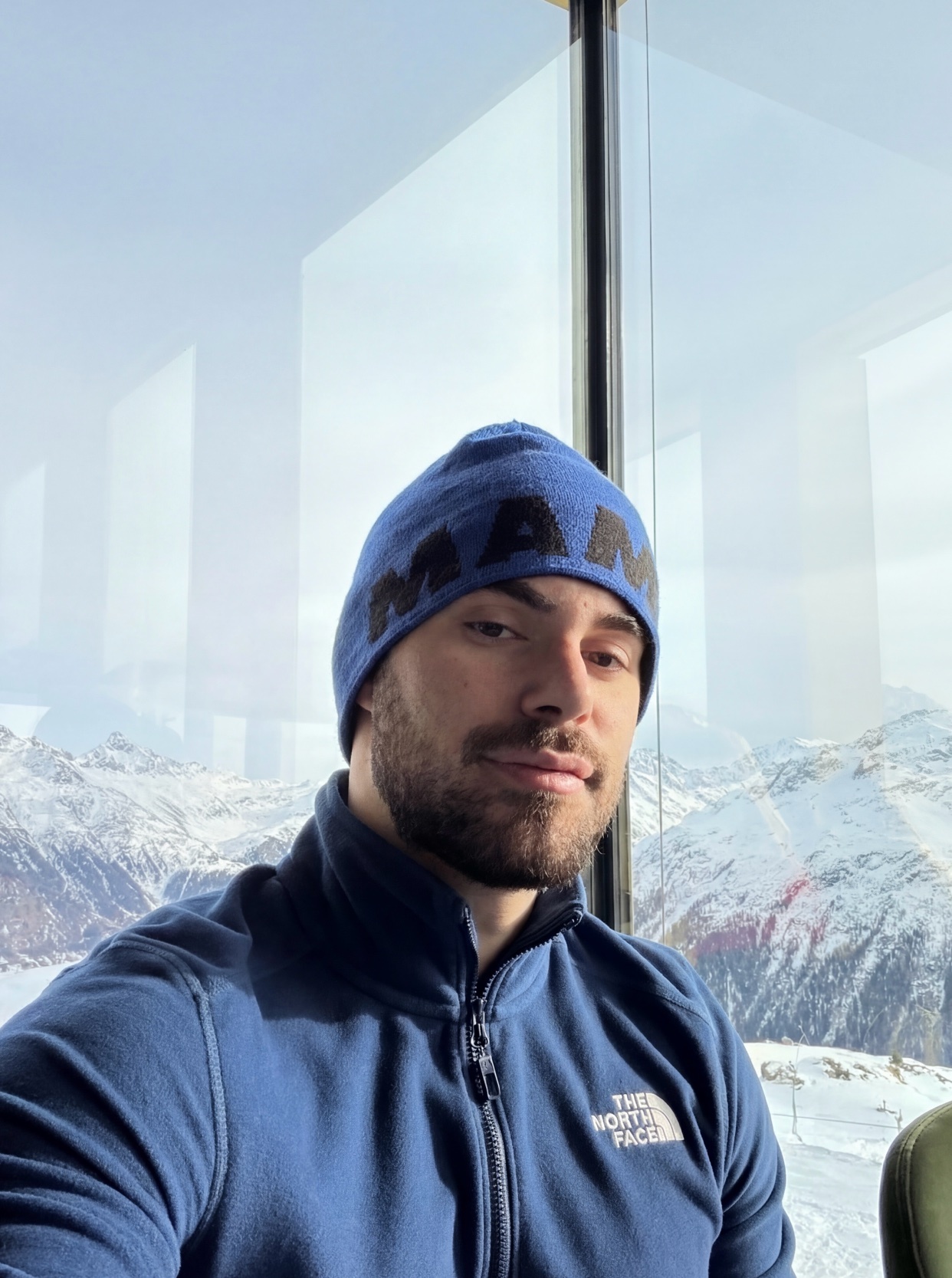 A man poses for a selfie in front of a window with a snowy mountain view. He is wearing a blue beanie and jacket.