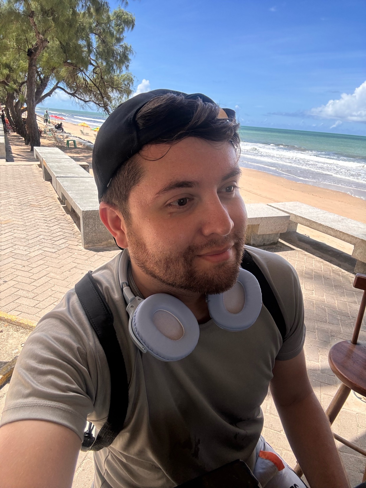 A man wearing a baseball cap and headphones poses for a selfie at the beach. He is wearing a grey t-shirt and has a backpack on.