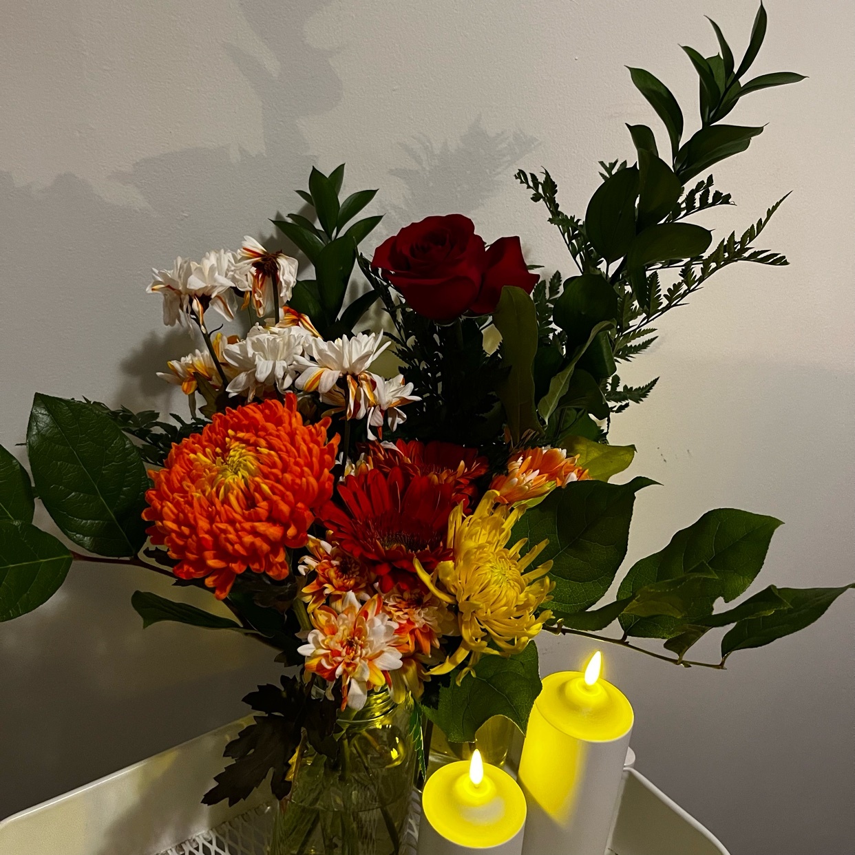 A beautiful bouquet of flowers with a red rose and orange and yellow flowers. Two candles are lit in front of the flowers.