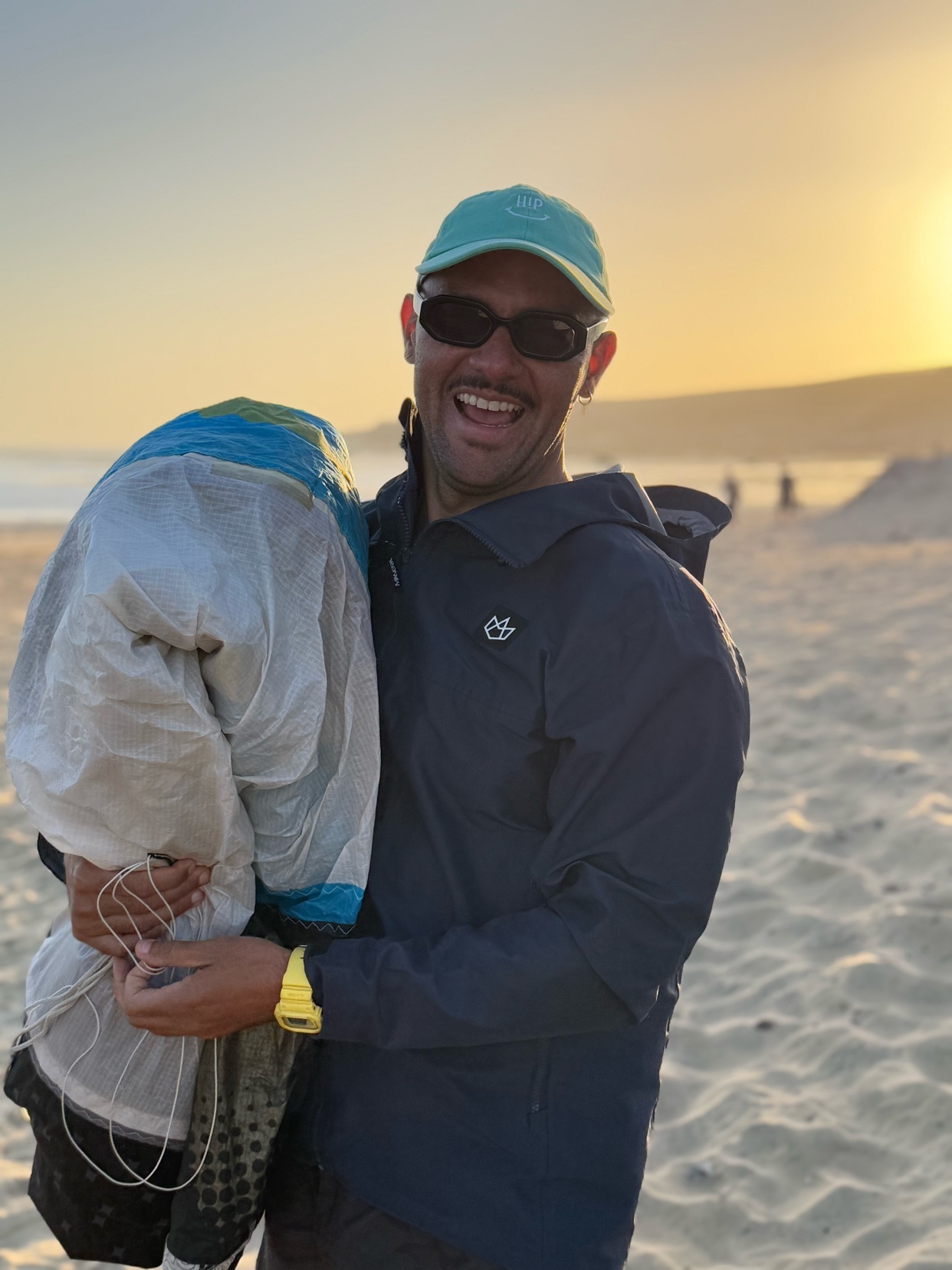 A man smiles while holding a kite on a beach. The sun sets in the background.
