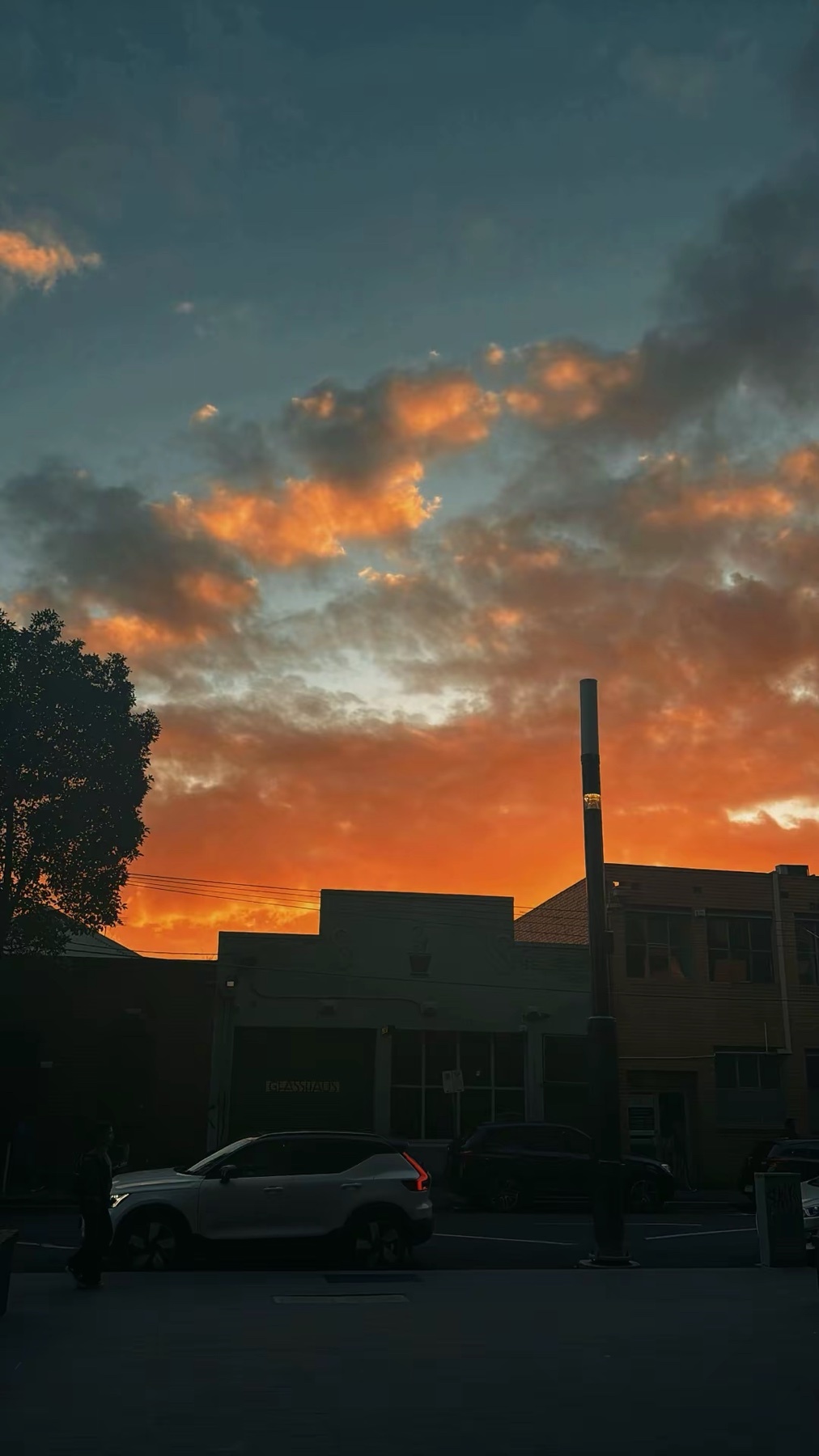 A dramatic sunset paints the sky with vibrant orange and yellow hues, set against a backdrop of dark, moody clouds. Silhouetted buildings and a street with cars create an urban evening scene.