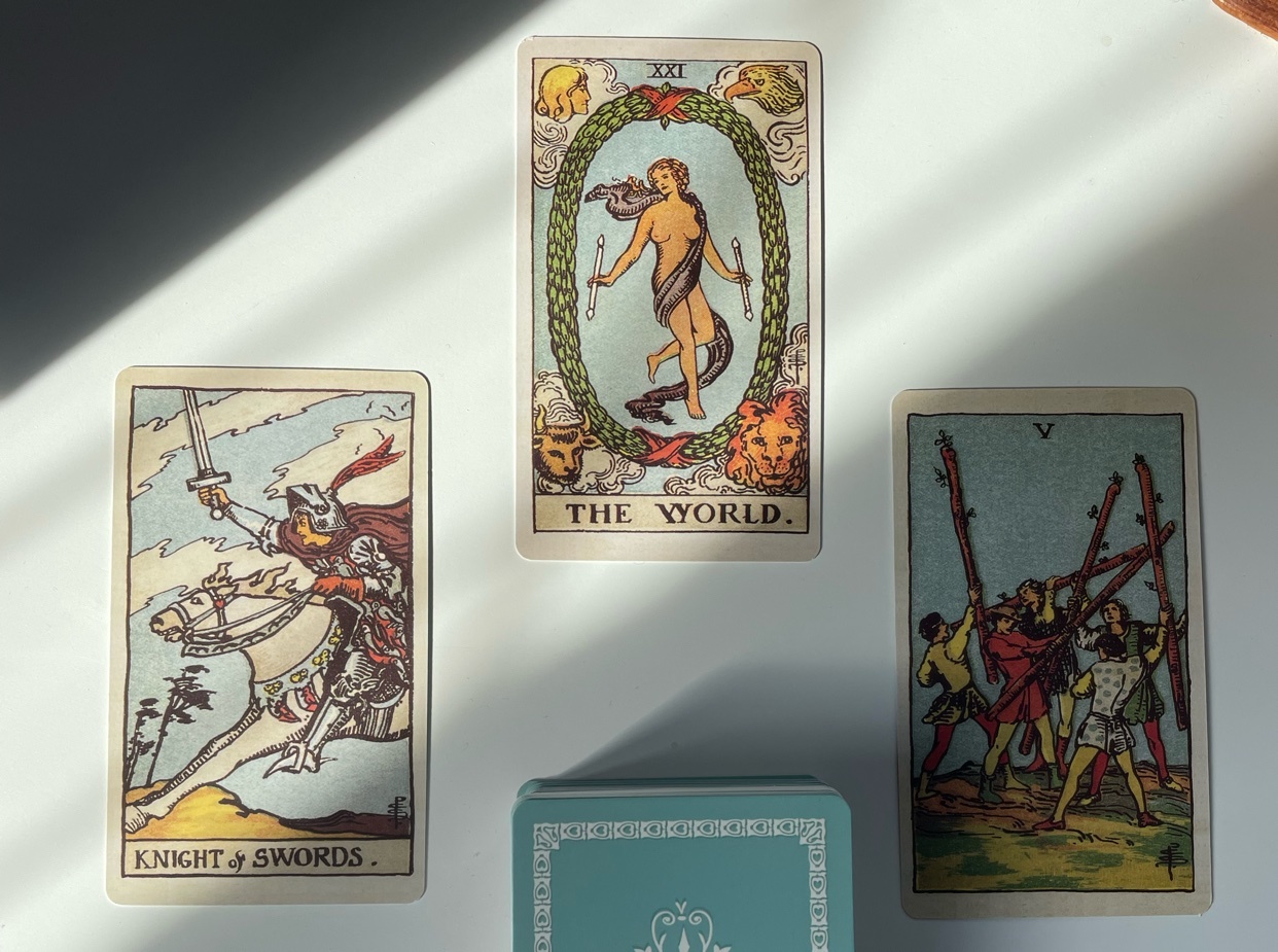 A close-up shot of a tarot card reading, featuring the cards "The World," "Knight of Swords," and "Five of Wands."