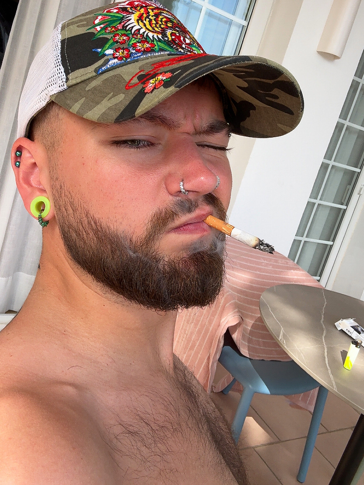 A man with a beard and piercings smokes a cigarette. He is shirtless and wearing a hat.