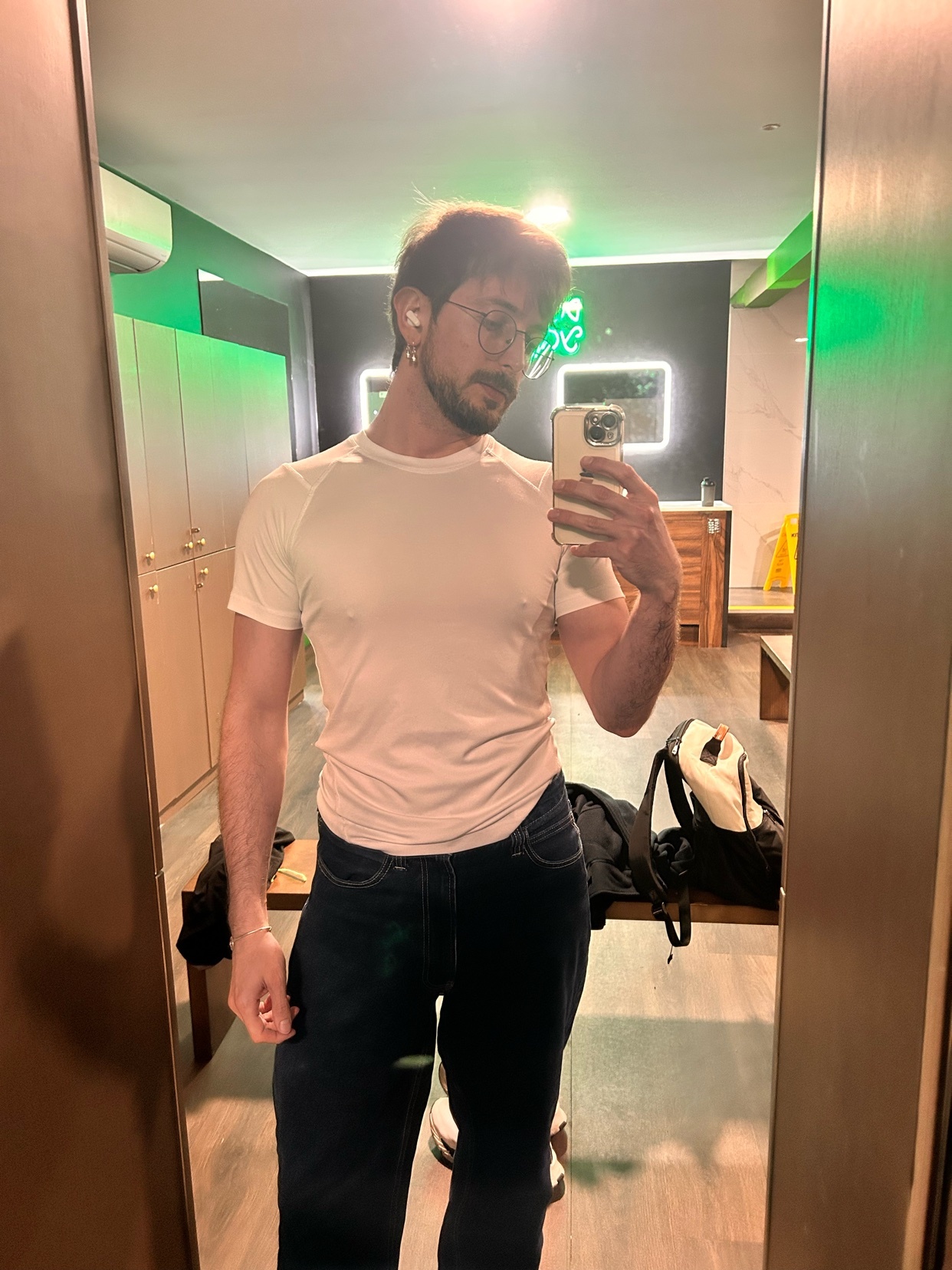 A man with glasses and a beard takes a mirror selfie in a gym locker room. He is wearing a white t-shirt and dark jeans.