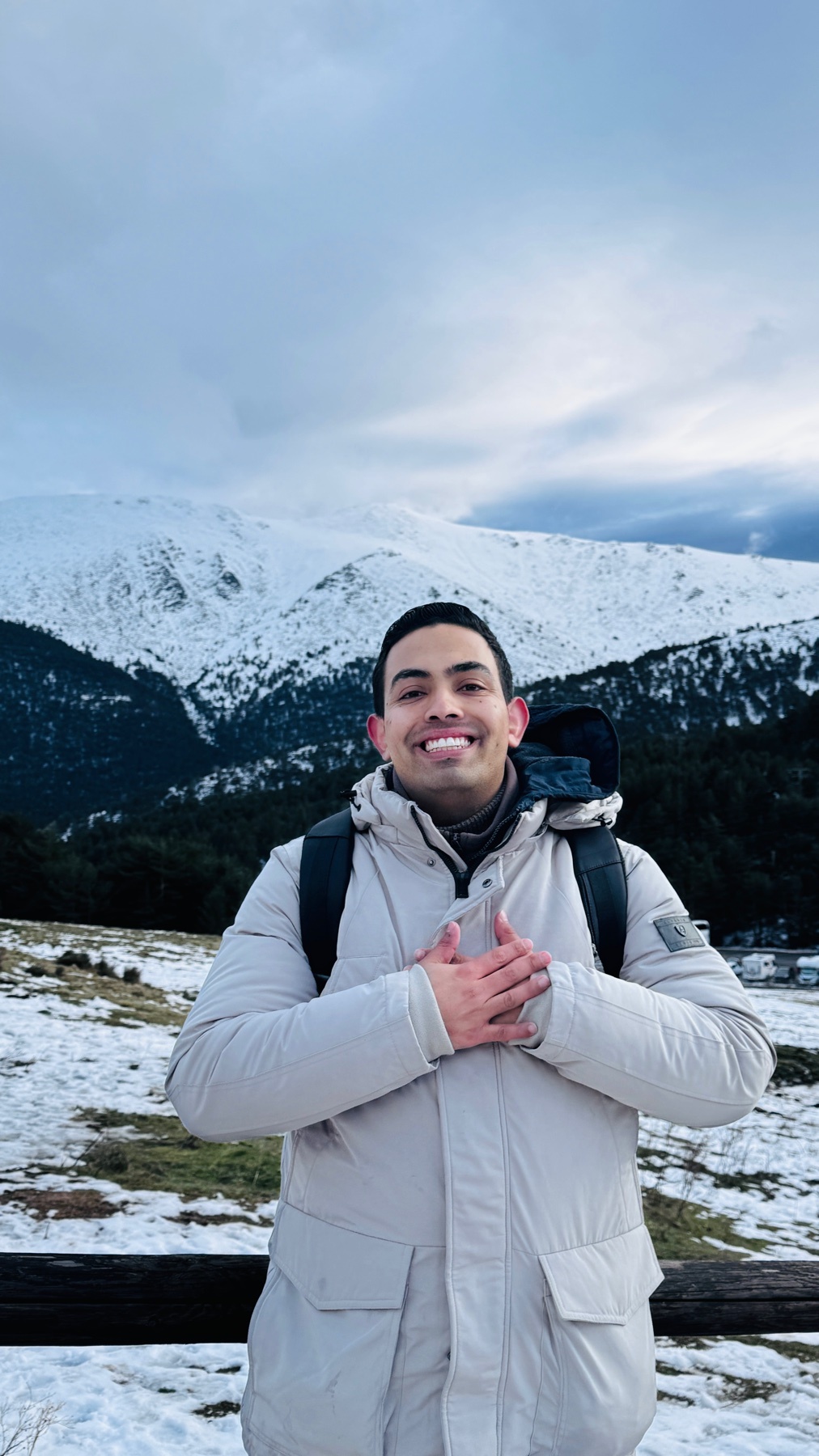 A man smiles in front of a snowy mountain range. He is wearing a winter jacket and backpack.