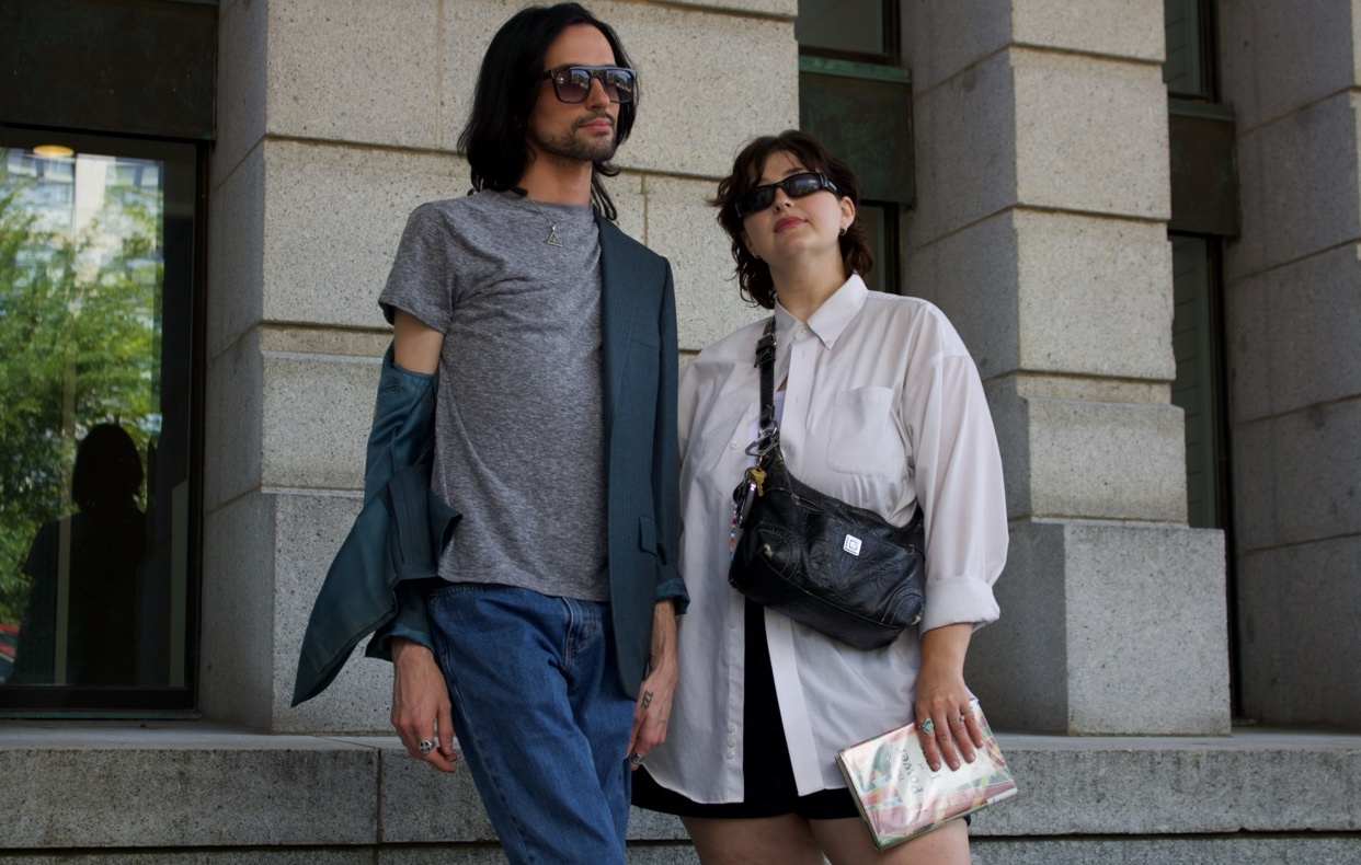 A man and a woman stand on steps outside a building, both wearing sunglasses. The man has long dark hair and is wearing a grey t-shirt, blue jeans, and a dark blazer. The woman has dark hair and is wearing a white oversized button-down shirt, black s