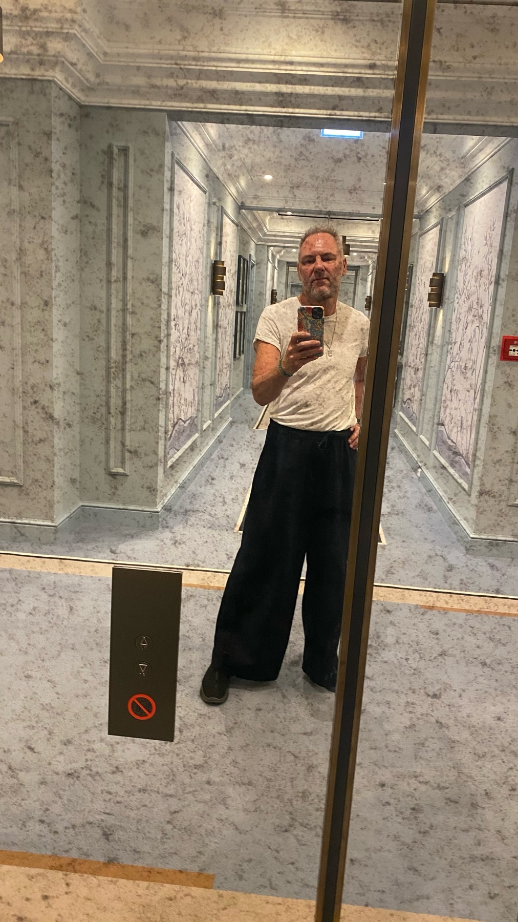 A man takes a selfie in an elevator, wearing a white t-shirt and dark, wide-leg pants. The elevator hallway has marble-patterned walls and ornate lighting fixtures.