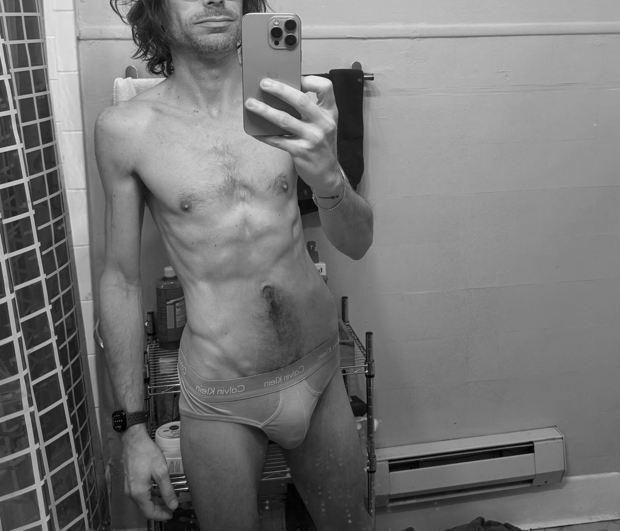 A black and white selfie of a man in Calvin Klein underwear in a bathroom. He has long hair and visible abs.