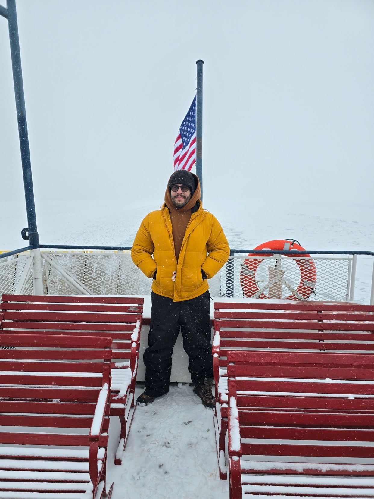 A man stands on a ferry deck in the snow, with rows of red seats and an American flag behind him.