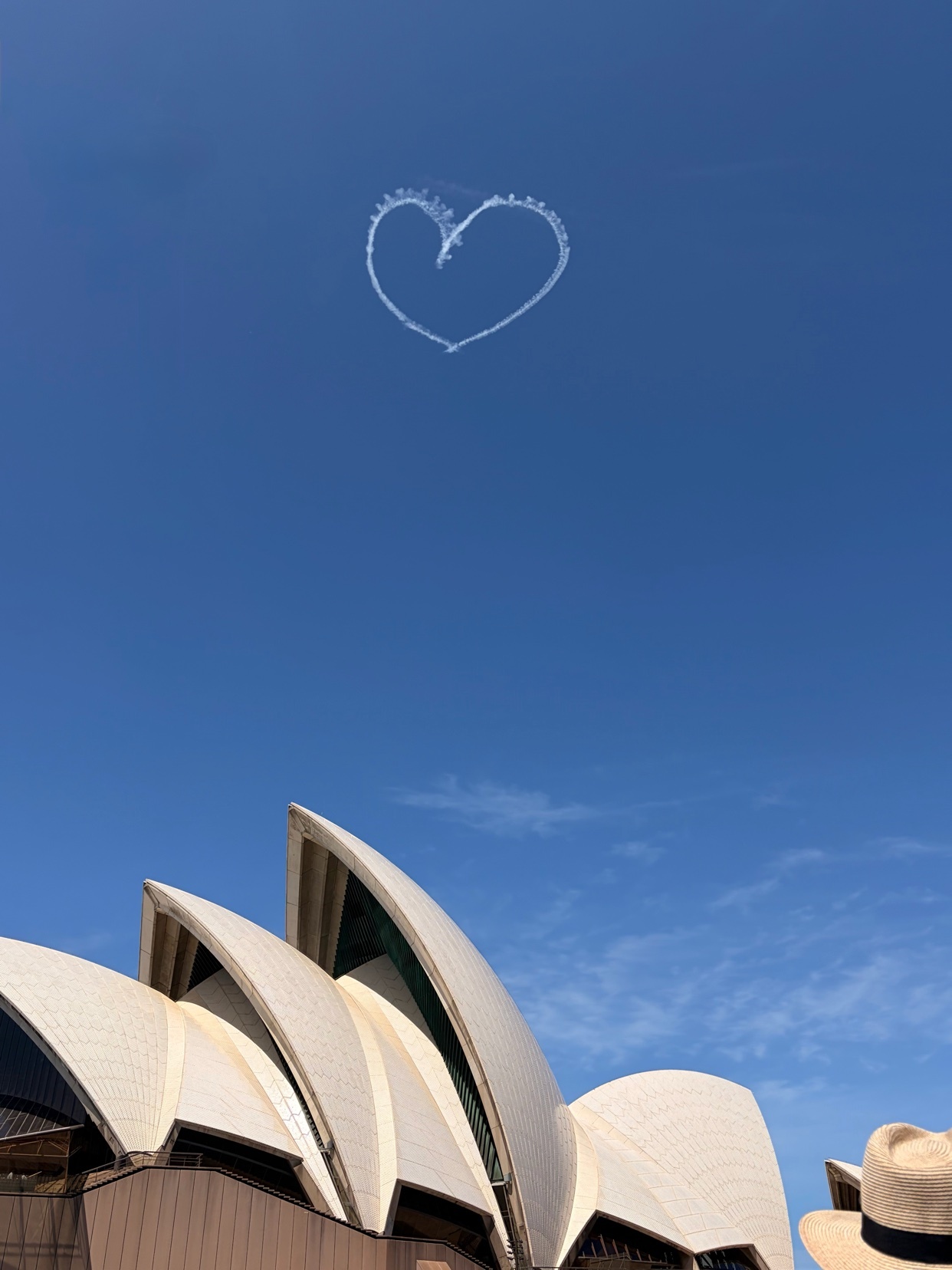 A heart shape is drawn in the sky with smoke above the iconic Sydney Opera House on a clear day.