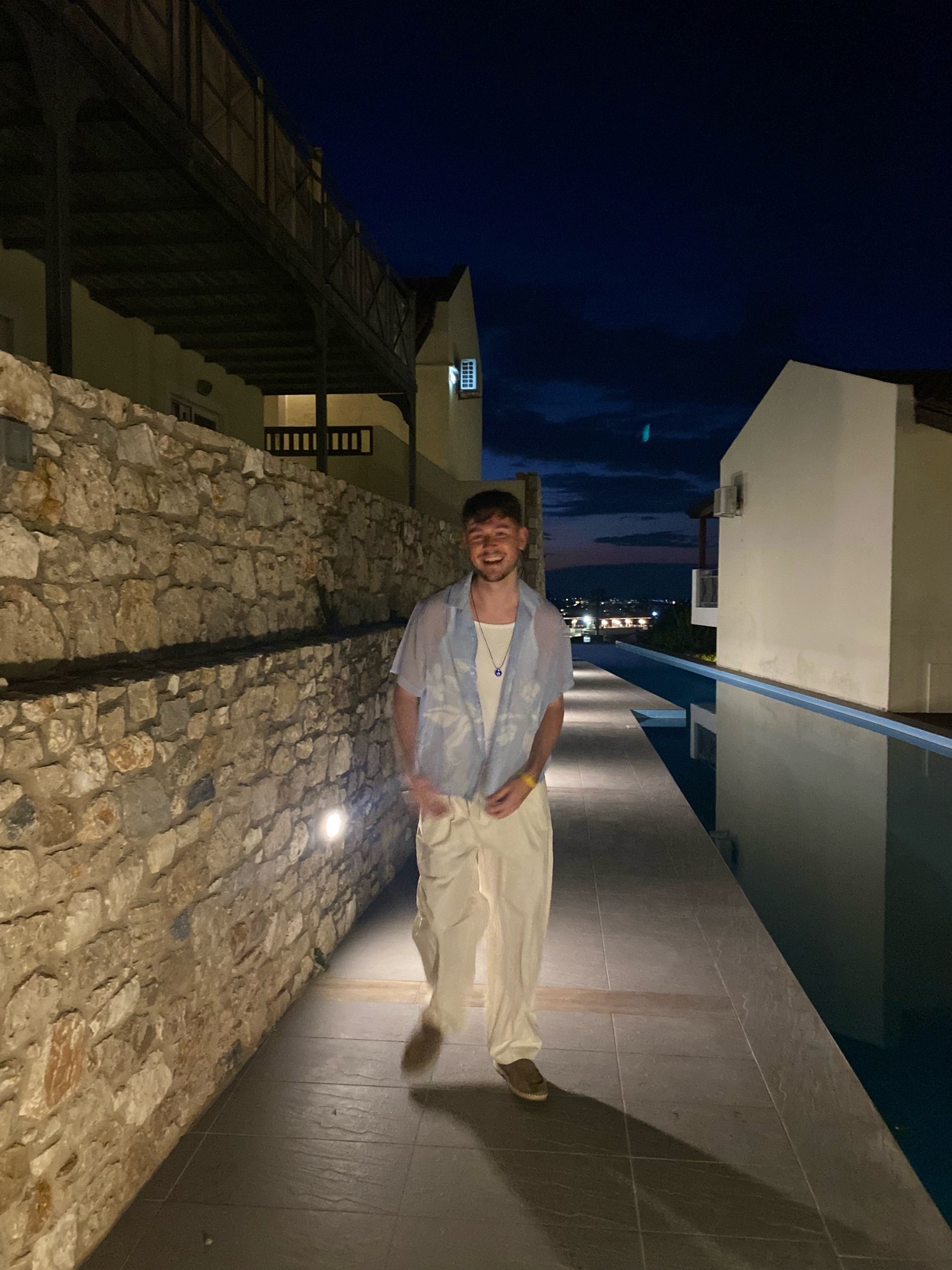 A man poses for a photo at night, wearing a stylish outfit. The photo captures a modern aesthetic with architectural elements in the background.