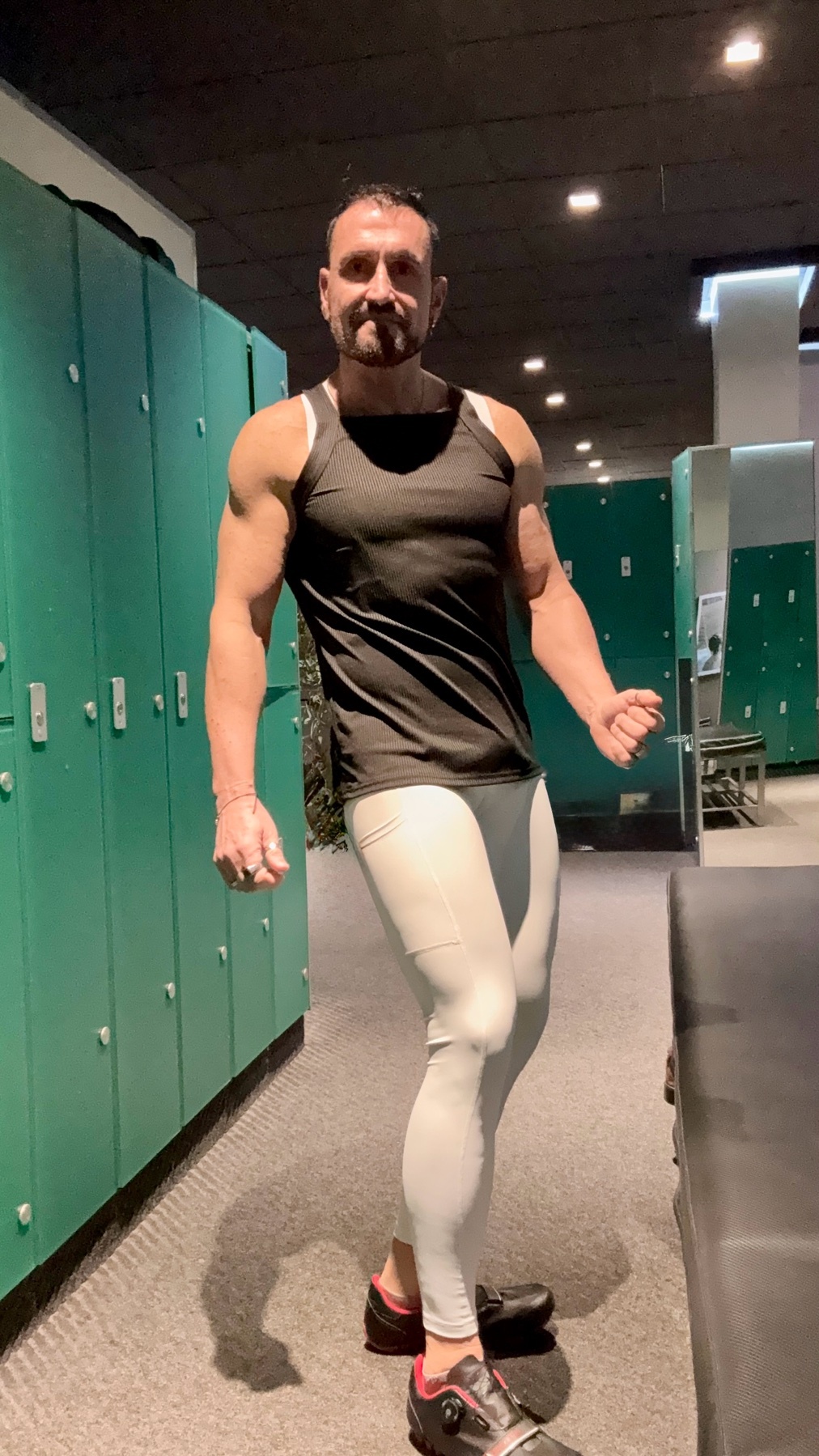 A muscular man in a black tank top and white leggings poses in a gym locker room.