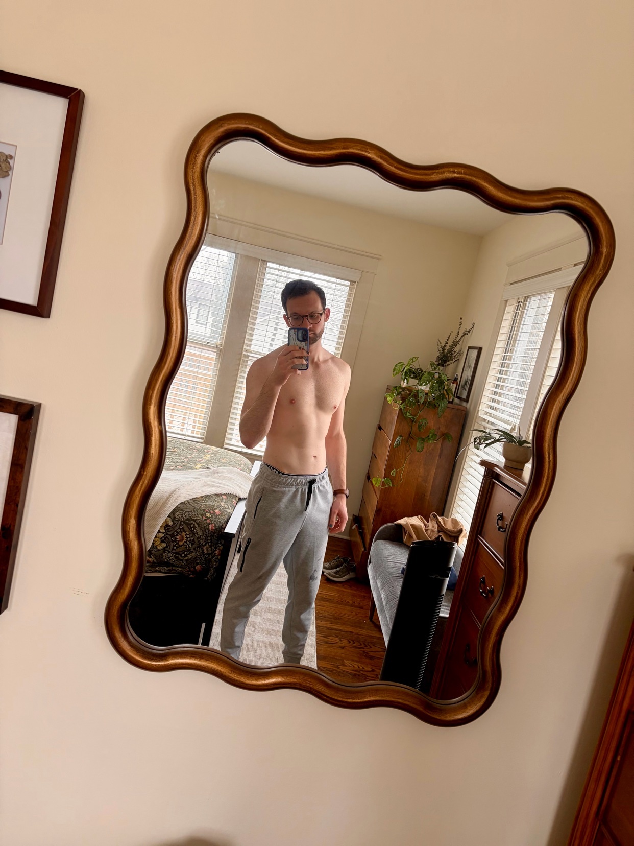 A man takes a mirror selfie, shirtless, in a bedroom. He is wearing grey sweatpants and glasses.