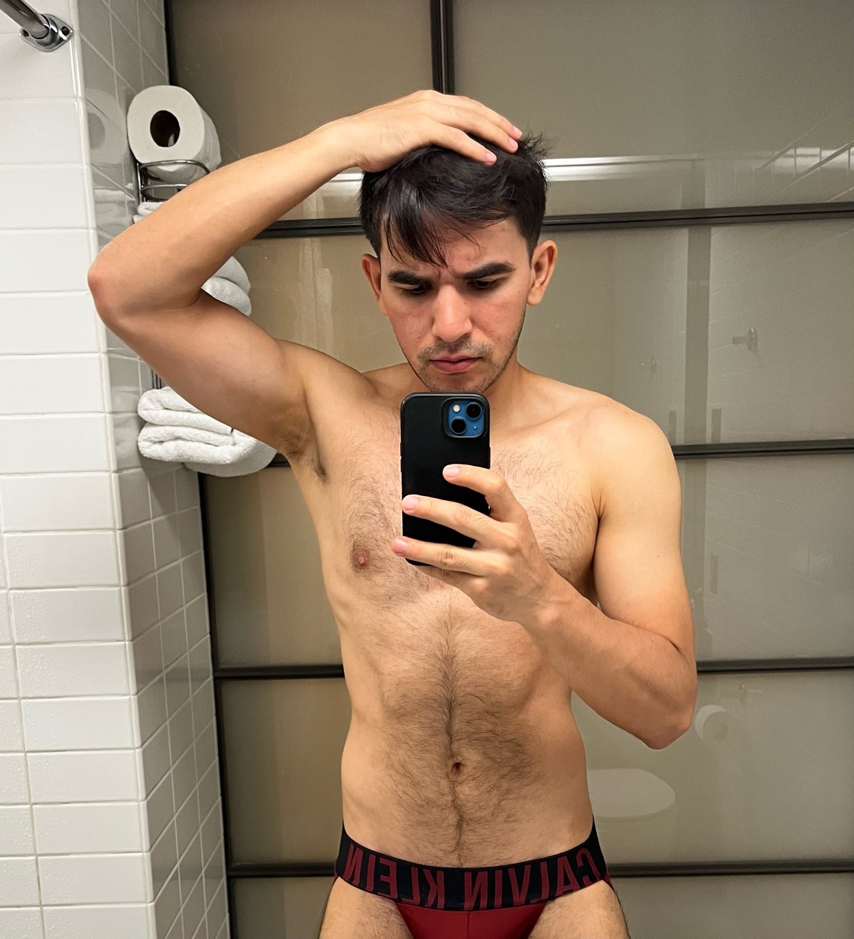 A man takes a selfie in a bathroom, wearing red Calvin Klein underwear. He is touching his hair with one hand and holding his phone with the other.