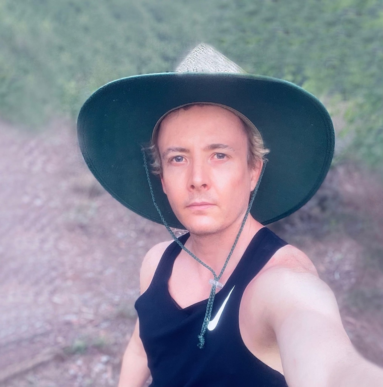 A man wearing a wide-brimmed hat and a black tank top takes a selfie outdoors.
