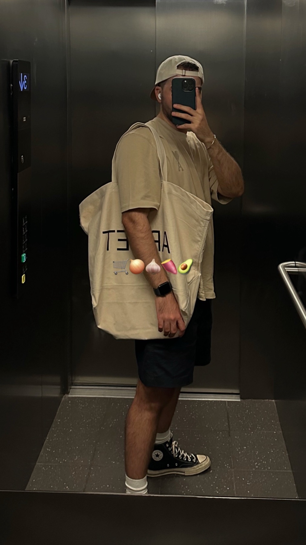 A man takes a mirror selfie in an elevator, holding a large tote bag. The bag has food emojis on it.