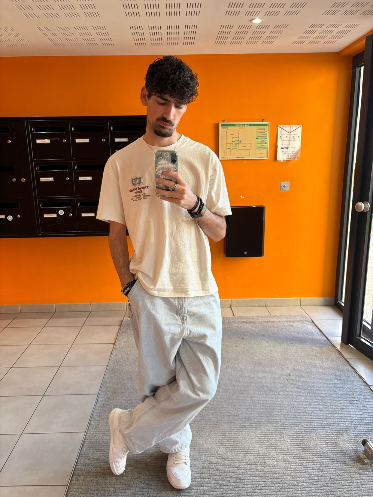 A man takes a selfie in a hallway with orange walls. He is wearing a cream-colored t-shirt and light blue baggy pants.