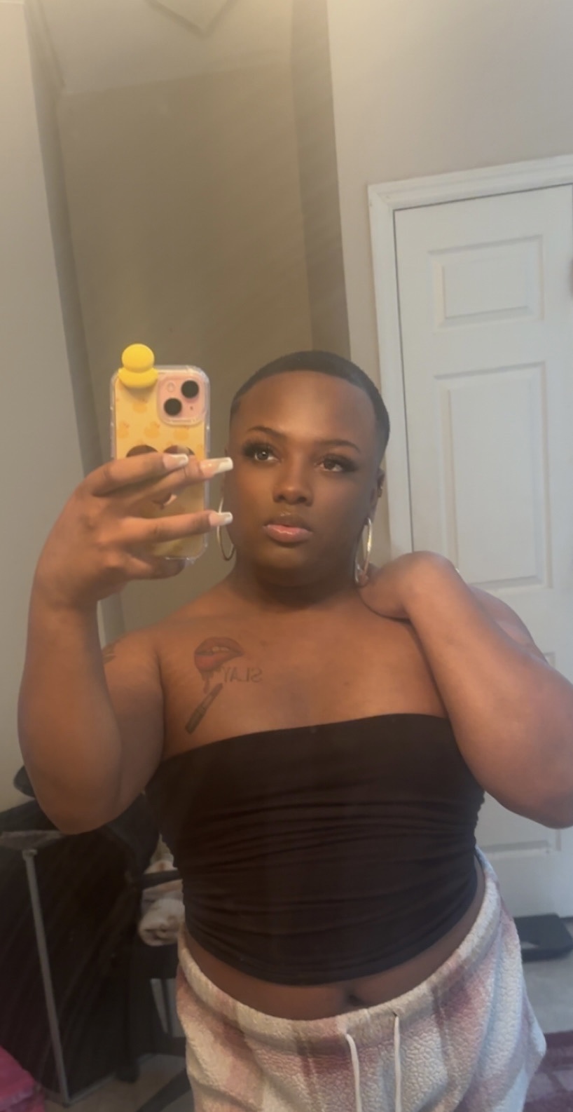 A person takes a mirror selfie, showcasing their makeup and strapless top. The image highlights their style and identity.
