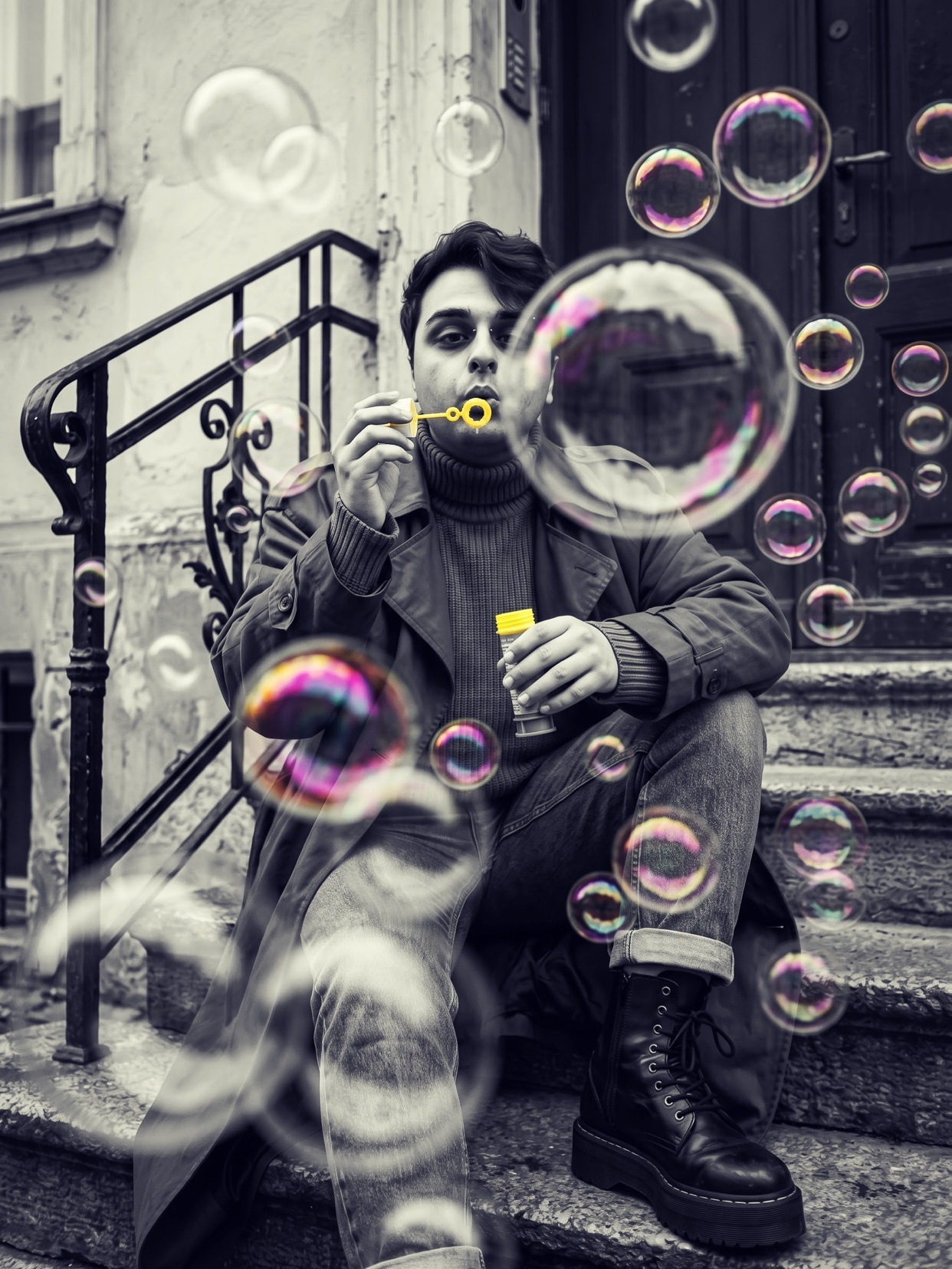 A man sits on outdoor stairs, blowing iridescent bubbles into the air. The black and white image is contrasted with the colorful bubbles, creating a whimsical and artistic effect.