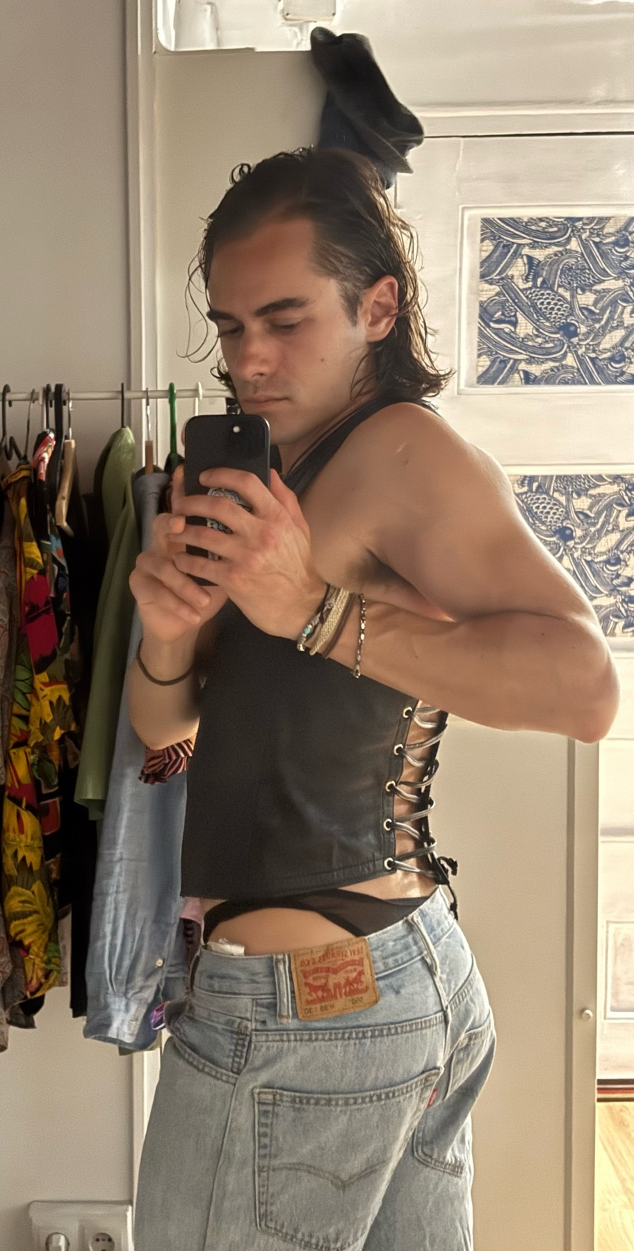 A man takes a mirror selfie, showing off his outfit. He is shirtless, wearing jeans and underwear.