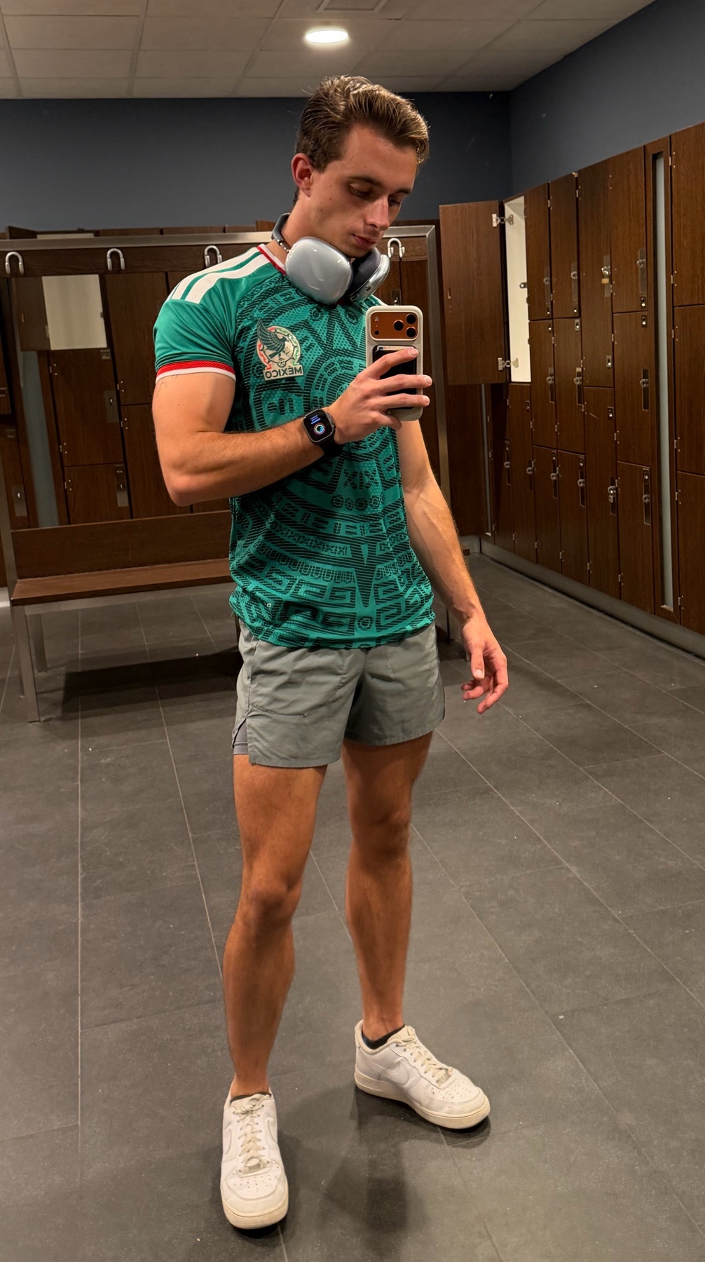 A man takes a selfie in a gym locker room, wearing a green Mexico jersey, grey shorts, and headphones around his neck.