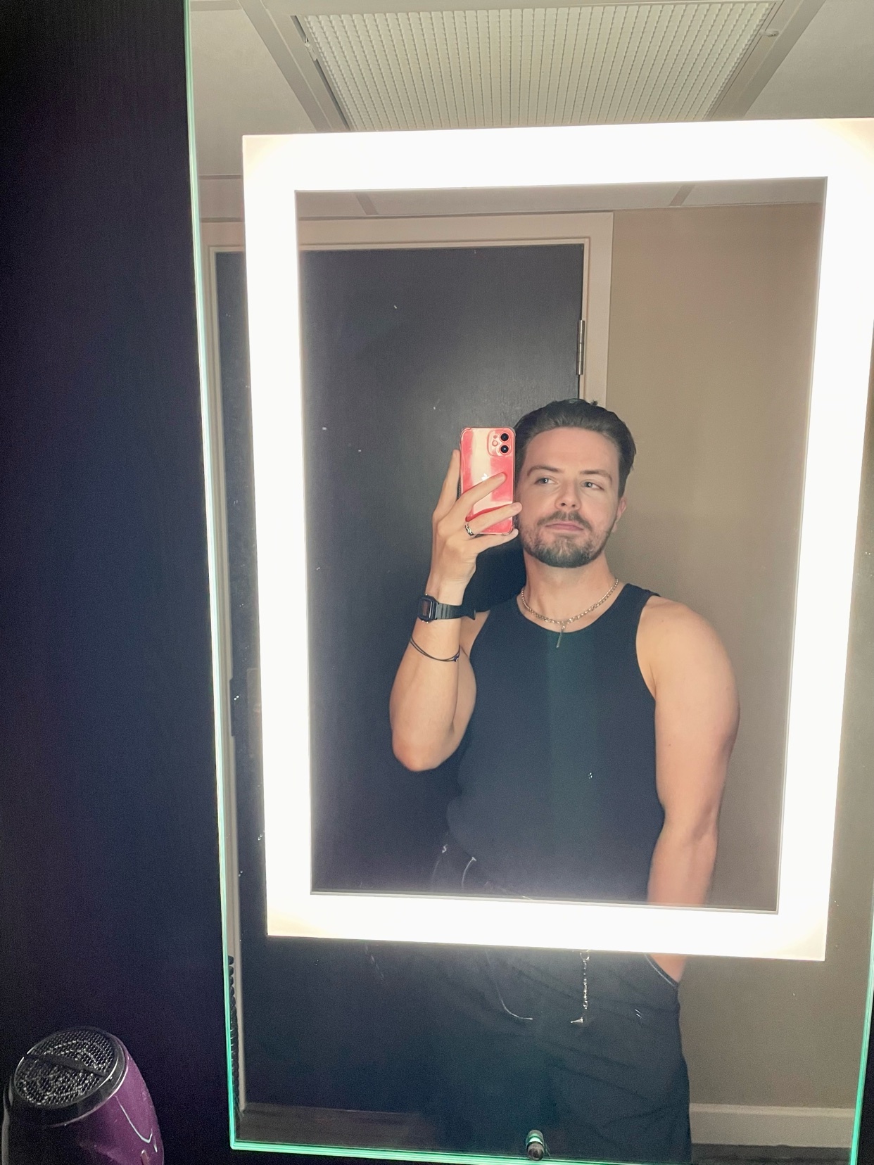 A man takes a mirror selfie, shirtless and flexing his arm. He is wearing a black tank top and black pants.