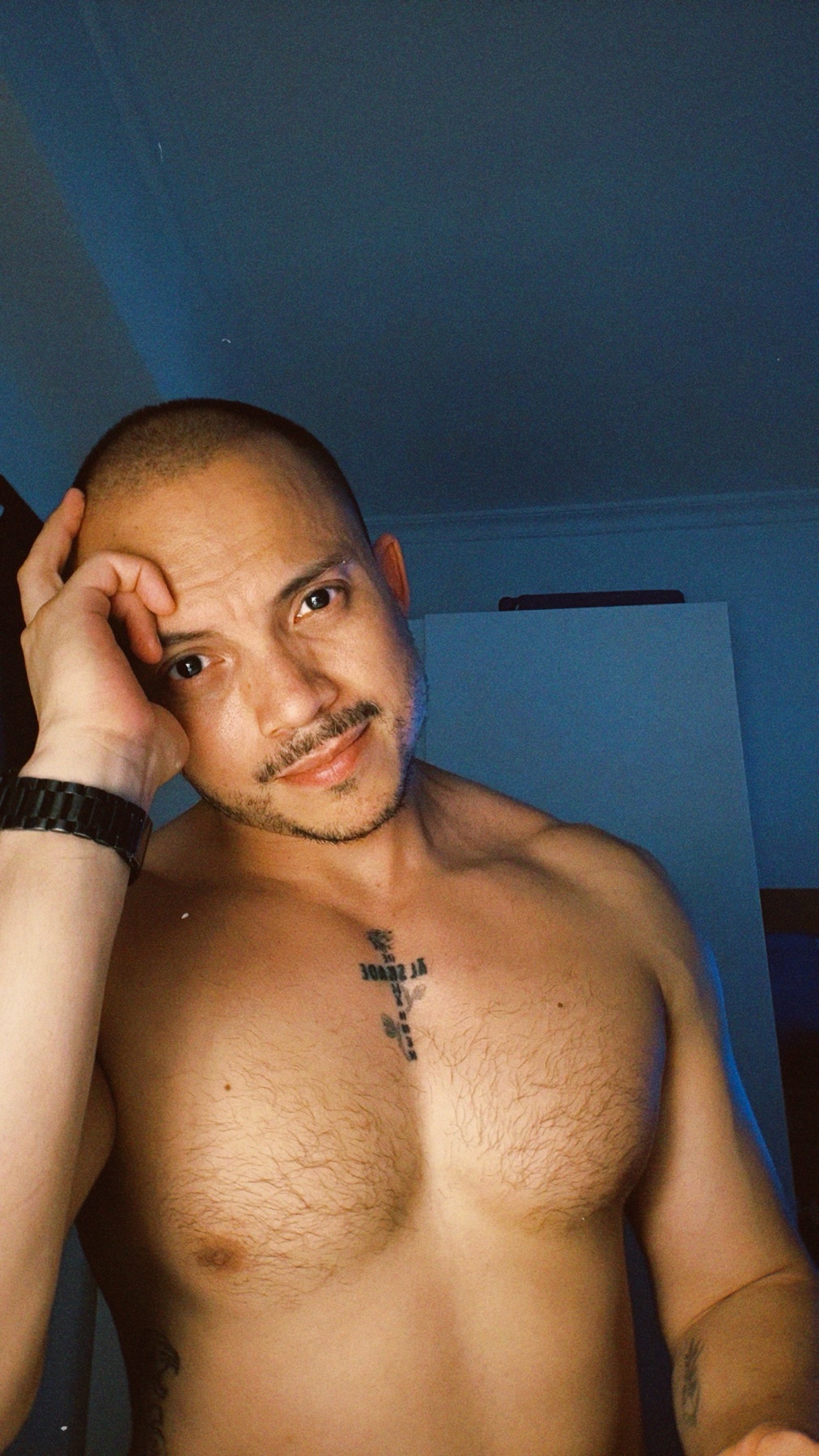 A man with a shaved head and stubble poses for a selfie, showing off his chest and arm tattoos. He is shirtless and looking towards the camera.