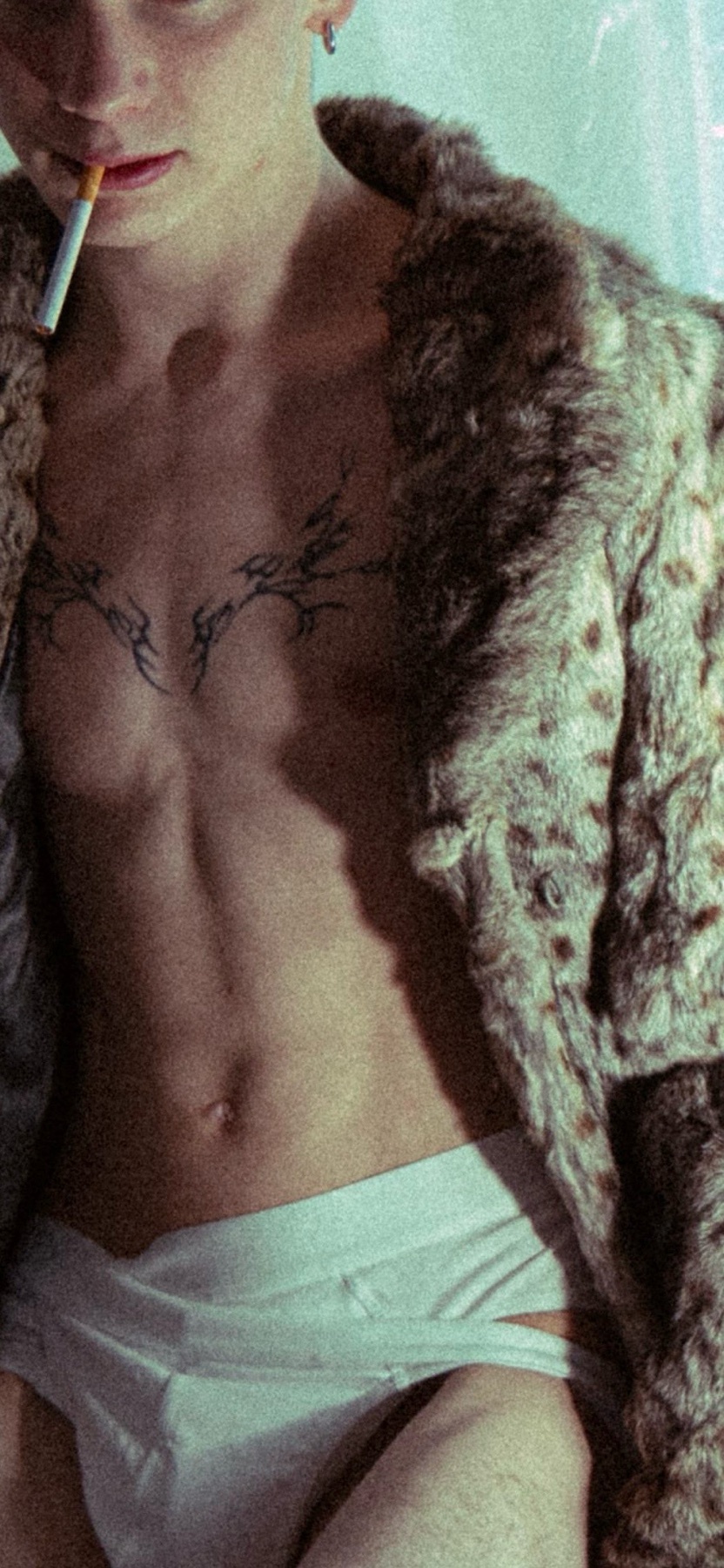 A man is seen shirtless, wearing underwear and a fur coat. He is smoking a cigarette and has a tattoo on his chest.