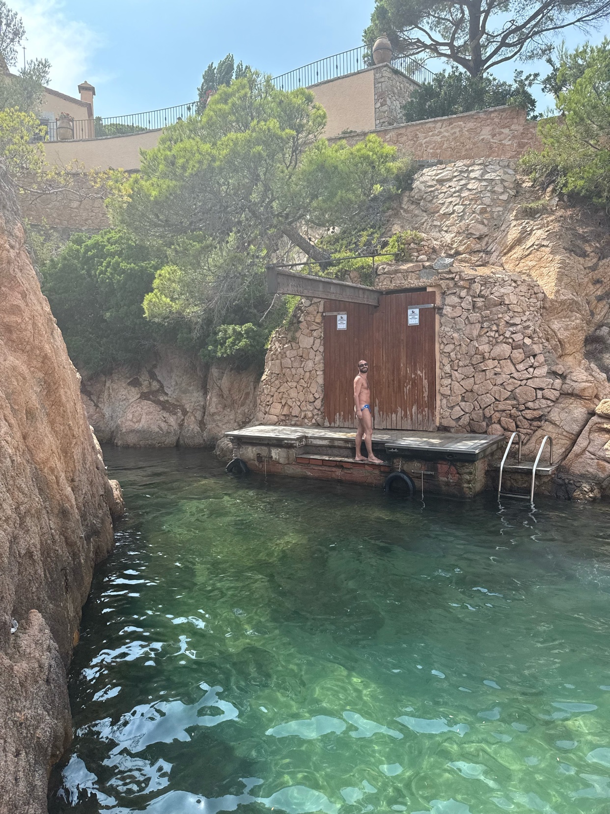 A woman in a bikini stands on a wooden dock next to a rustic wooden structure, with clear turquoise water lapping at the rocky shore. The scene evokes a longing for summer days and coastal relaxation.