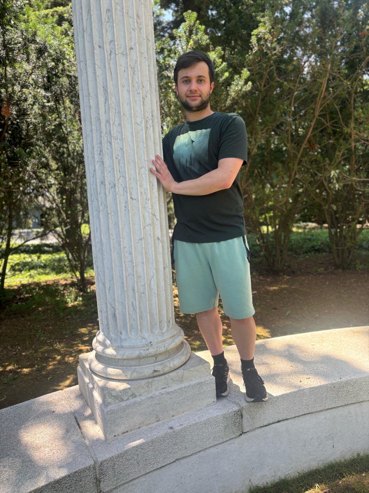 A man with stubble and dark hair stands outdoors next to a large marble column. He is wearing a dark green t-shirt and light green shorts, with black sneakers and socks.