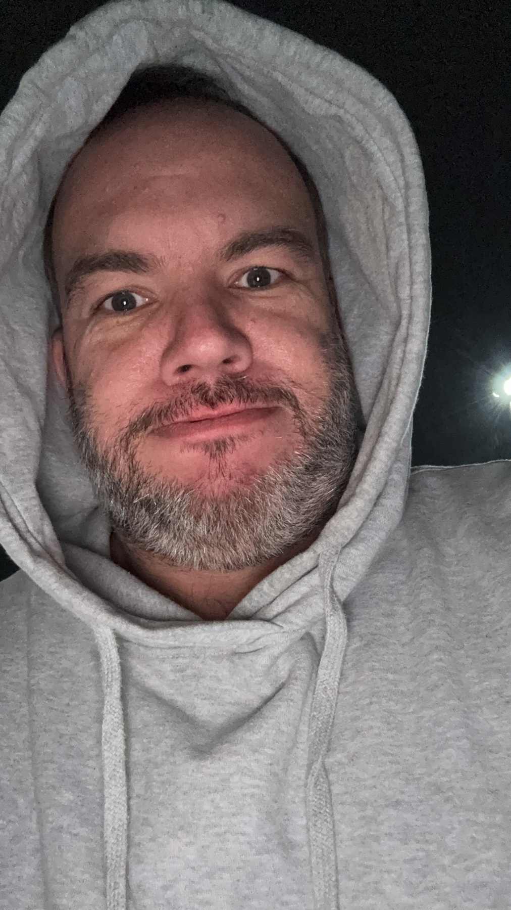 A man wearing a grey hoodie looks directly at the camera. The photo is a close-up portrait.