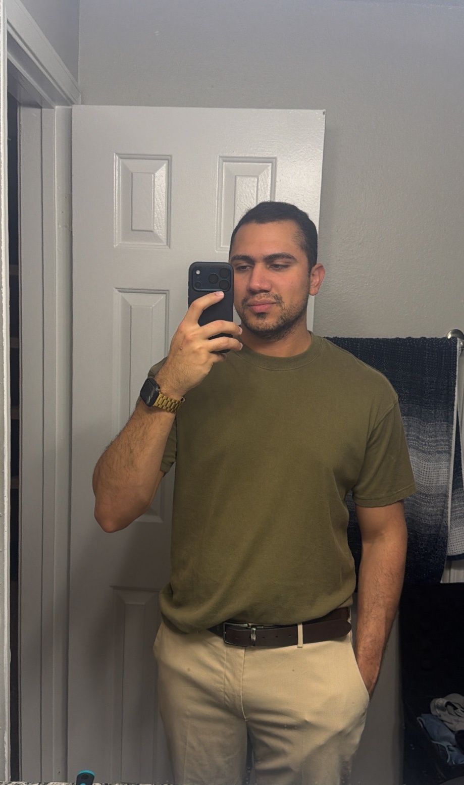 A man takes a mirror selfie in a bathroom. He is wearing a green t-shirt and khaki pants.