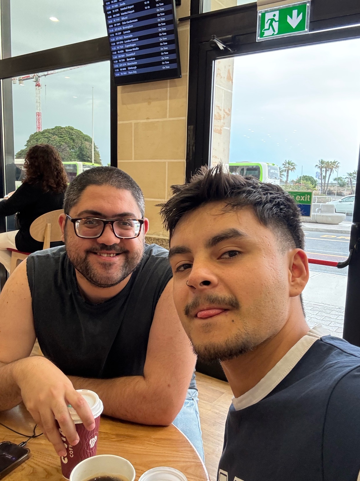 Two men smile for a selfie at a table with coffee cups. One man wears glasses and a dark t-shirt, while the other has stubble and a mustache and sticks his tongue out playfully.