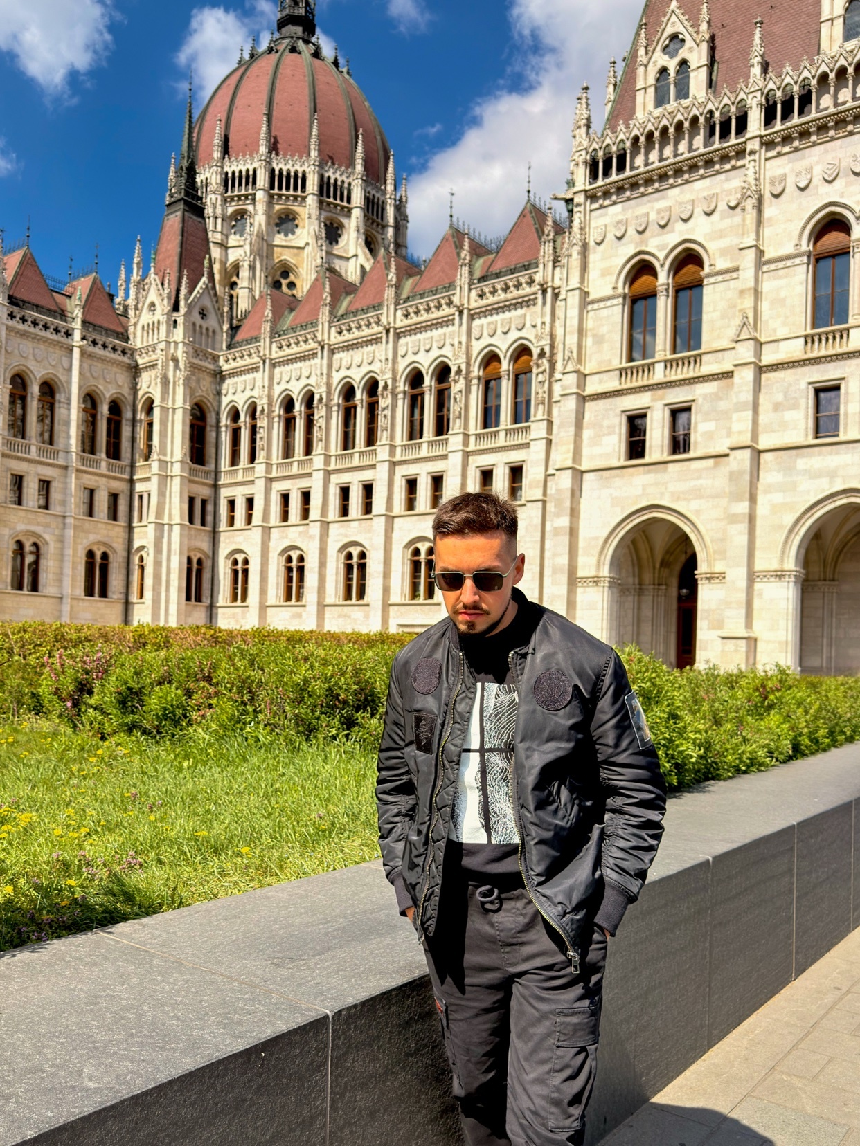 A man poses in front of the Hungarian Parliament Building in Budapest. He is wearing a black bomber jacket and sunglasses.