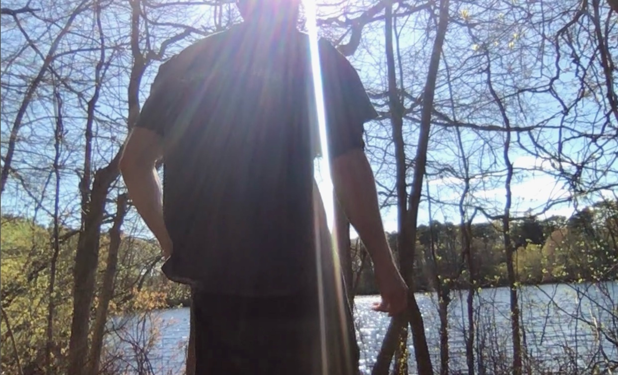 A person stands in a wooded area with a lake in the background. The sun is shining brightly, creating a lens flare effect.