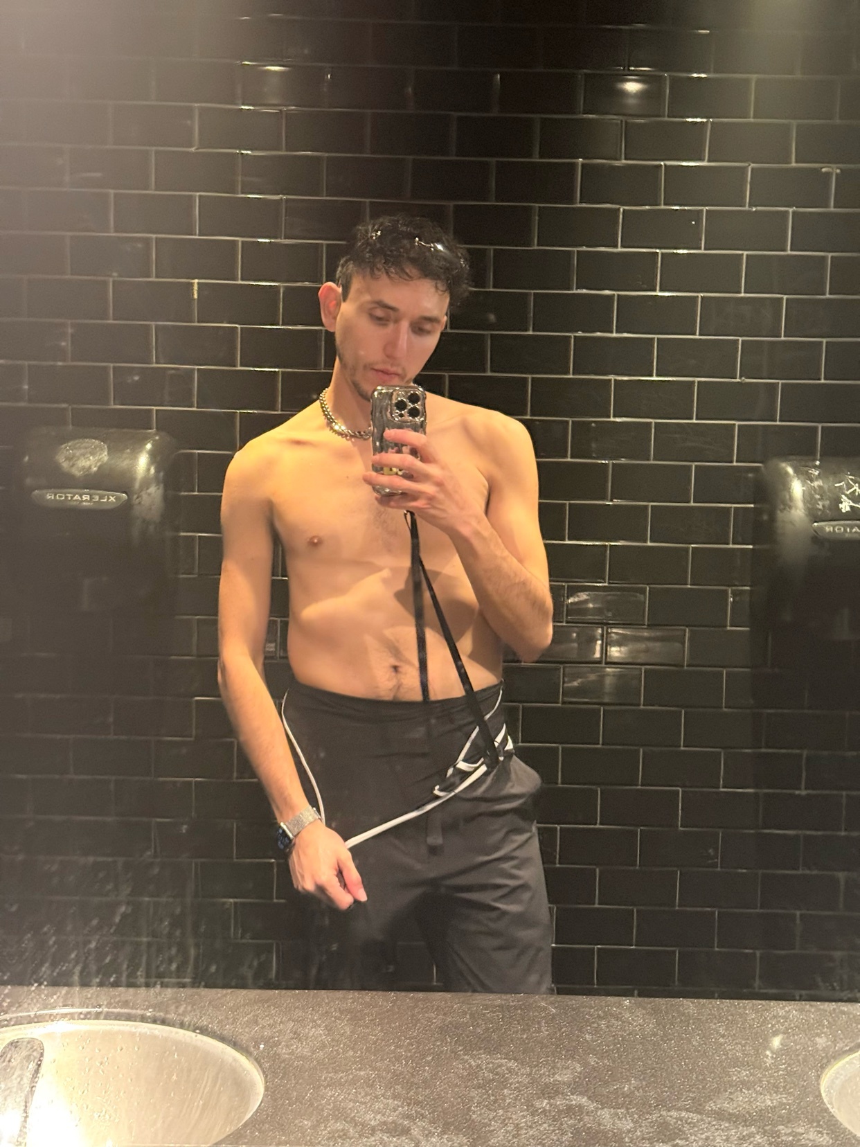 A man takes a shirtless mirror selfie in a bathroom. He is wearing black sweatpants and a silver necklace.