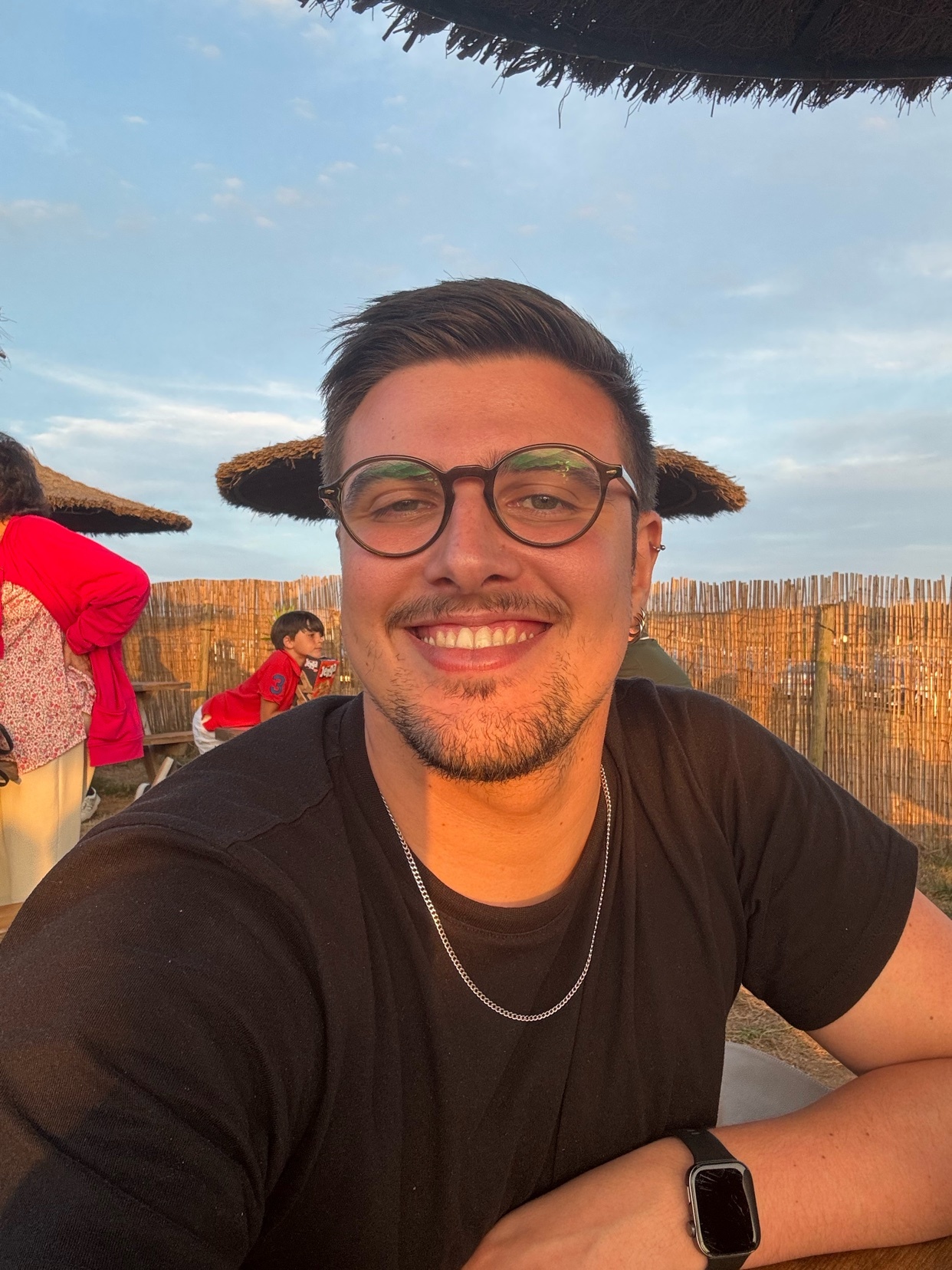 A man with glasses and stubble smiles at the camera during golden hour in Asturias. He is wearing a black t-shirt and a silver chain.