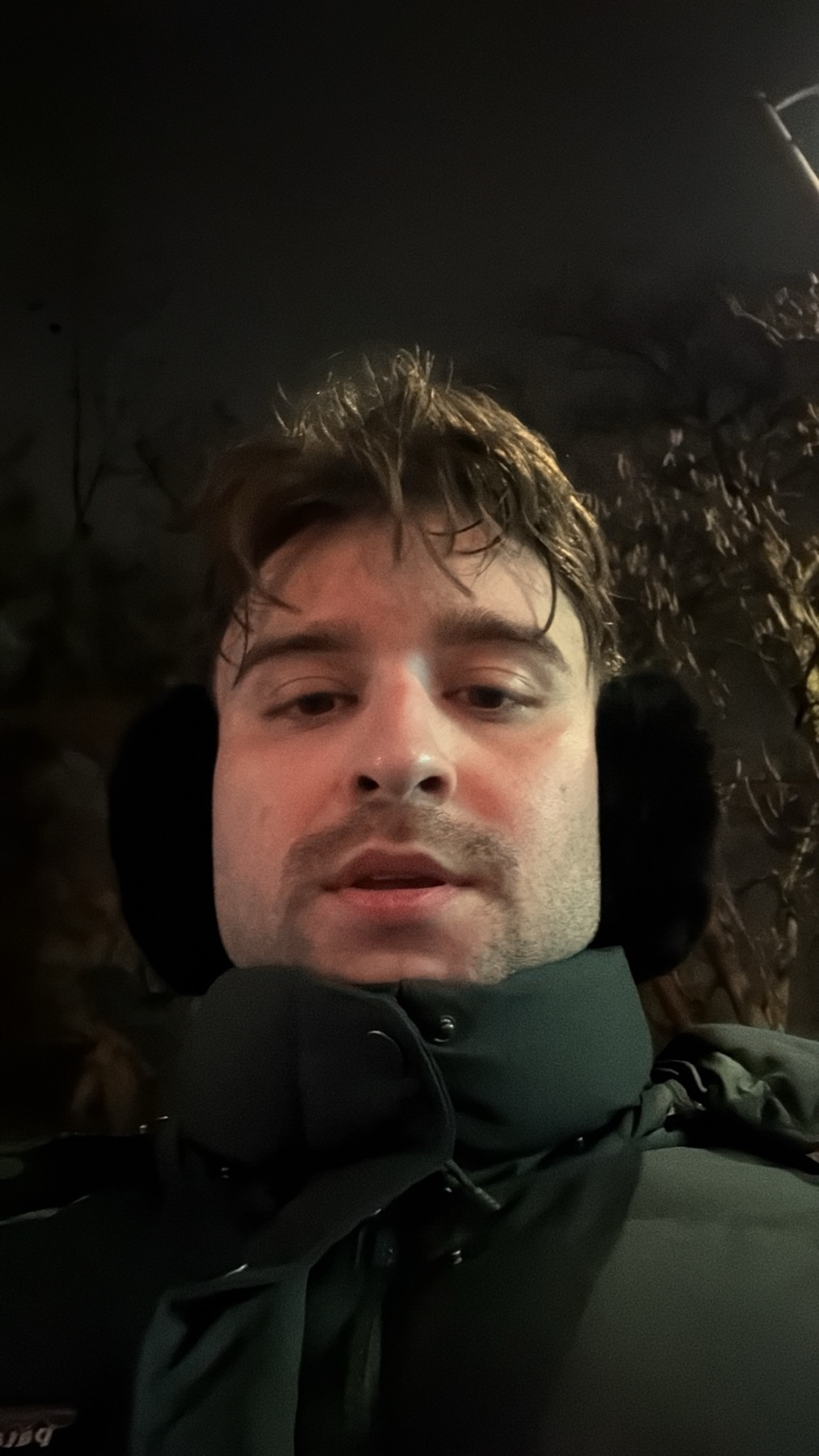A man takes a selfie outdoors at night. He is wearing earmuffs and a winter coat.